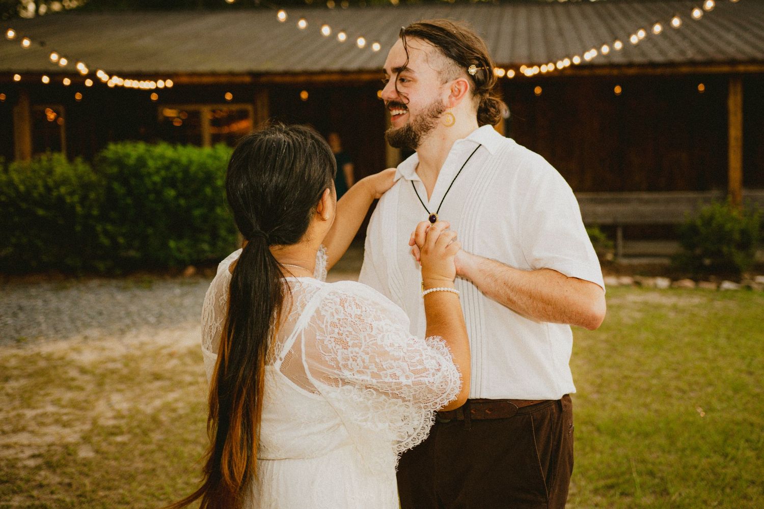 Bride in white lace dress adjusting groom's collar outdoors near a rustic barn with string lights.