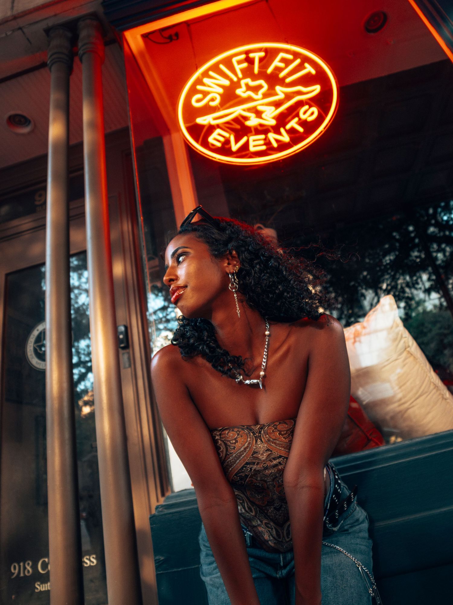 Fashionable Black woman leans forward under a red neon Swift Fit Events sign on a city street at dusk.