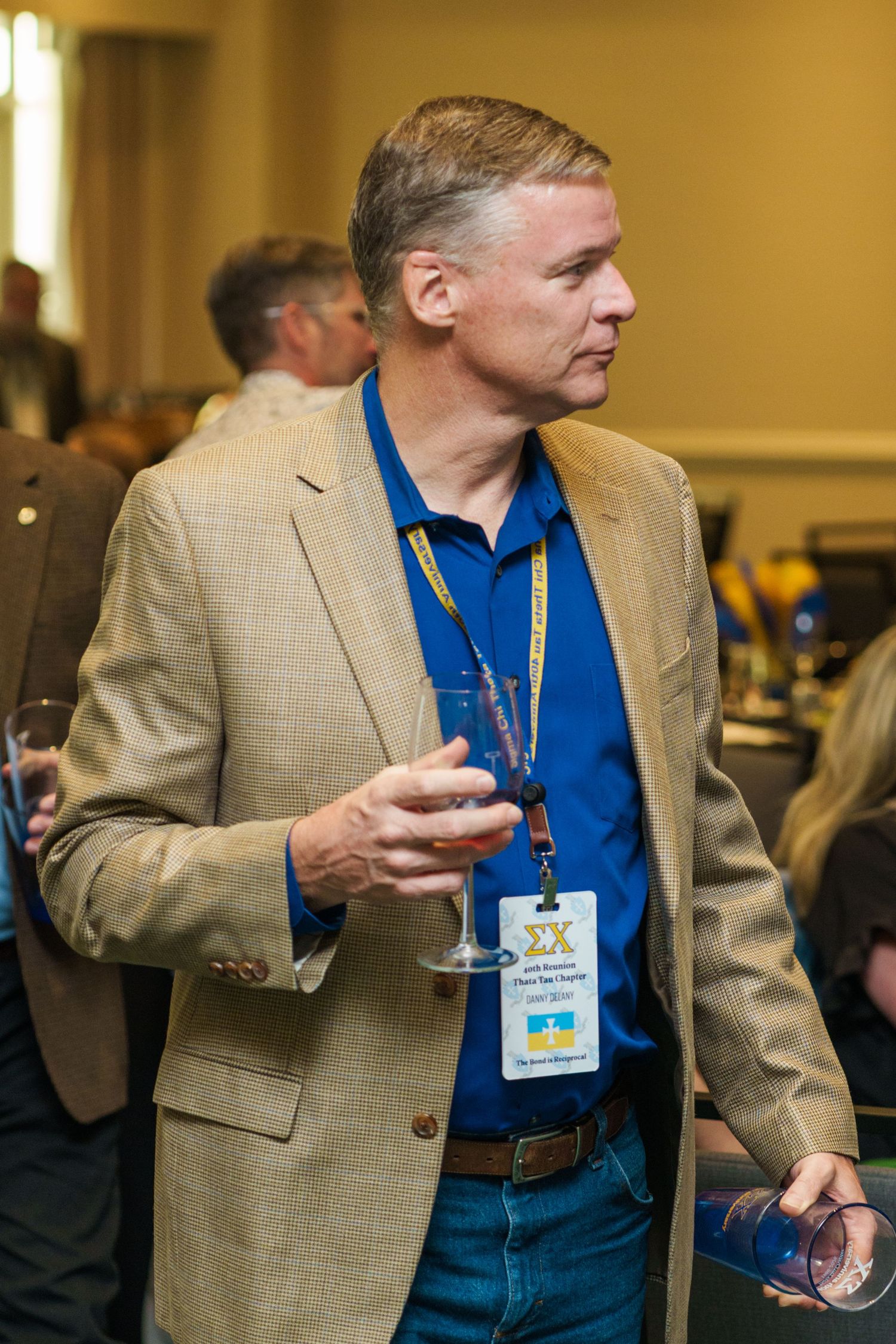 Man in blue shirt and tan blazer holds wine glass at networking event, wearing Sigma Chi lanyard badge.