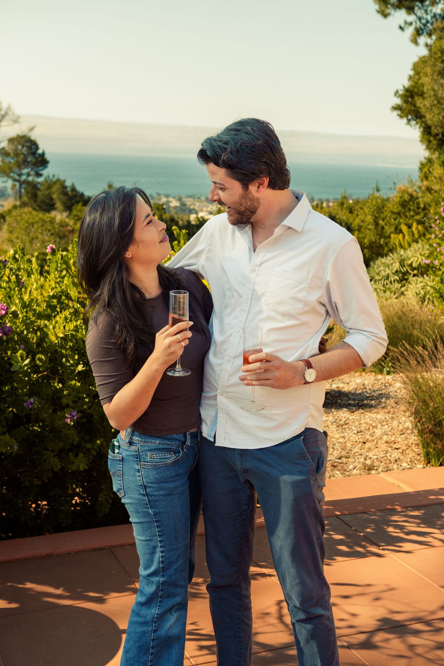 Couple smiling at each other while holding champagne glasses outdoors with ocean view and lush greenery in background.