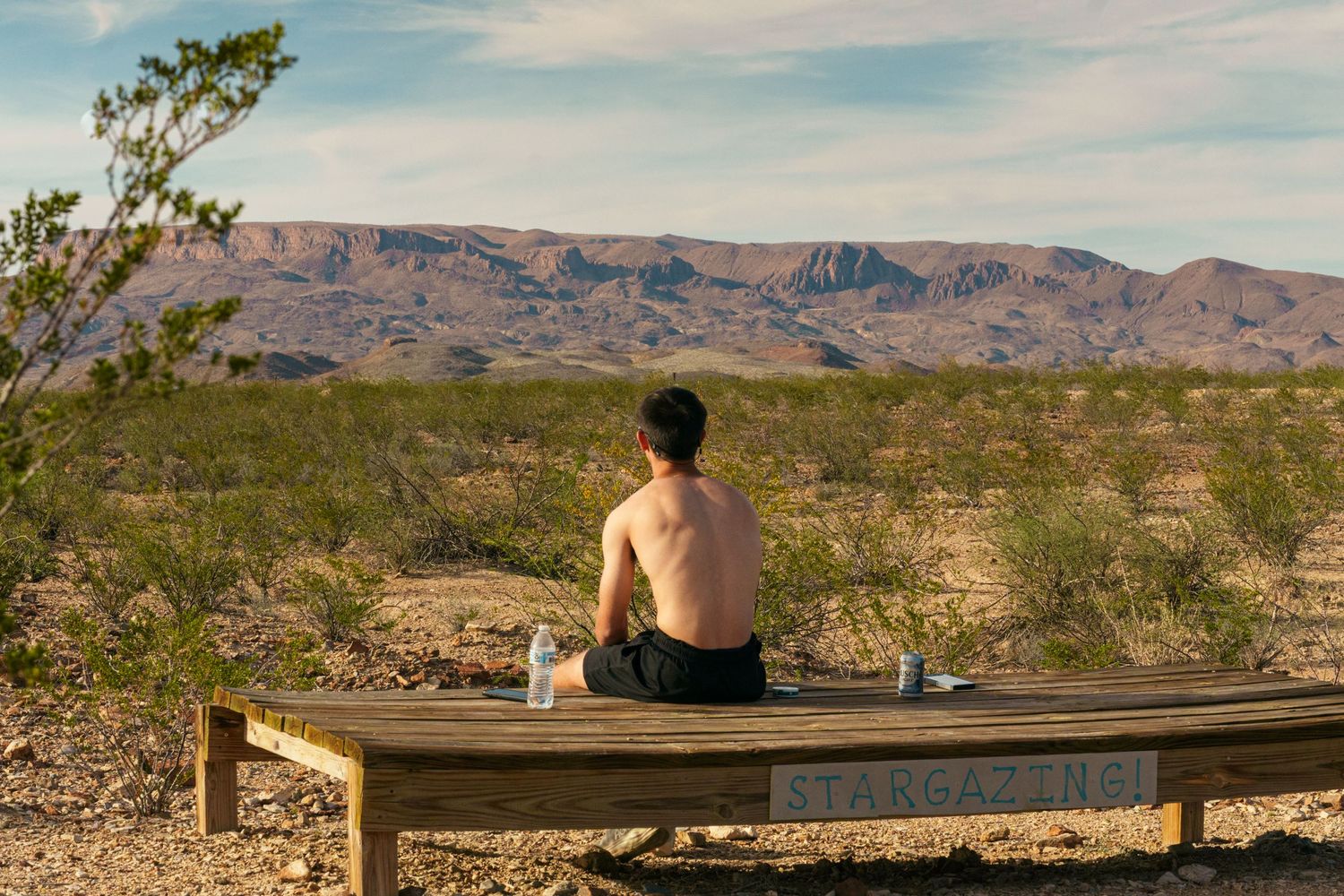 Shirtless man sitting on wooden deck gazing at desert mountain landscape in arid wilderness.
