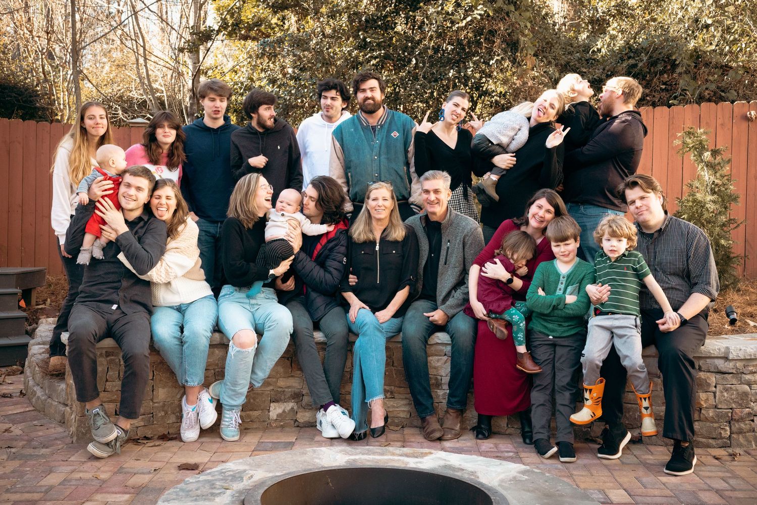 Large multigenerational family smiling together outdoors around a backyard fire pit.
