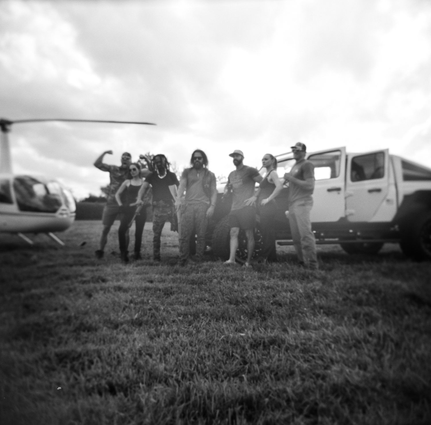 A film crew poses beside a helicopter and white SUV on a grass field in black and white.