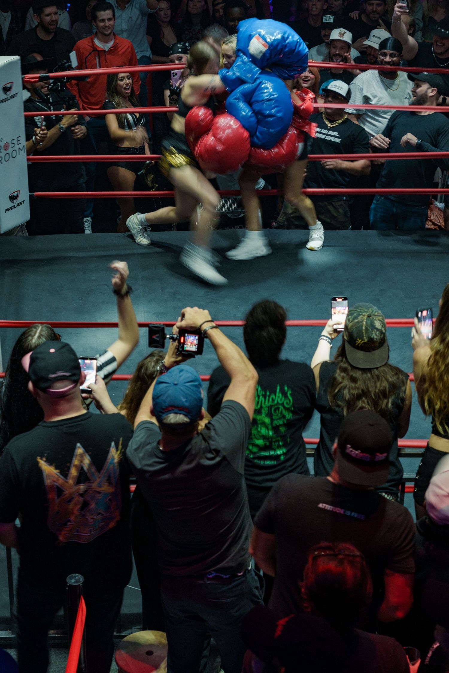 Audience members photograph two female boxers in oversized gloves competing in a lively boxing ring event.