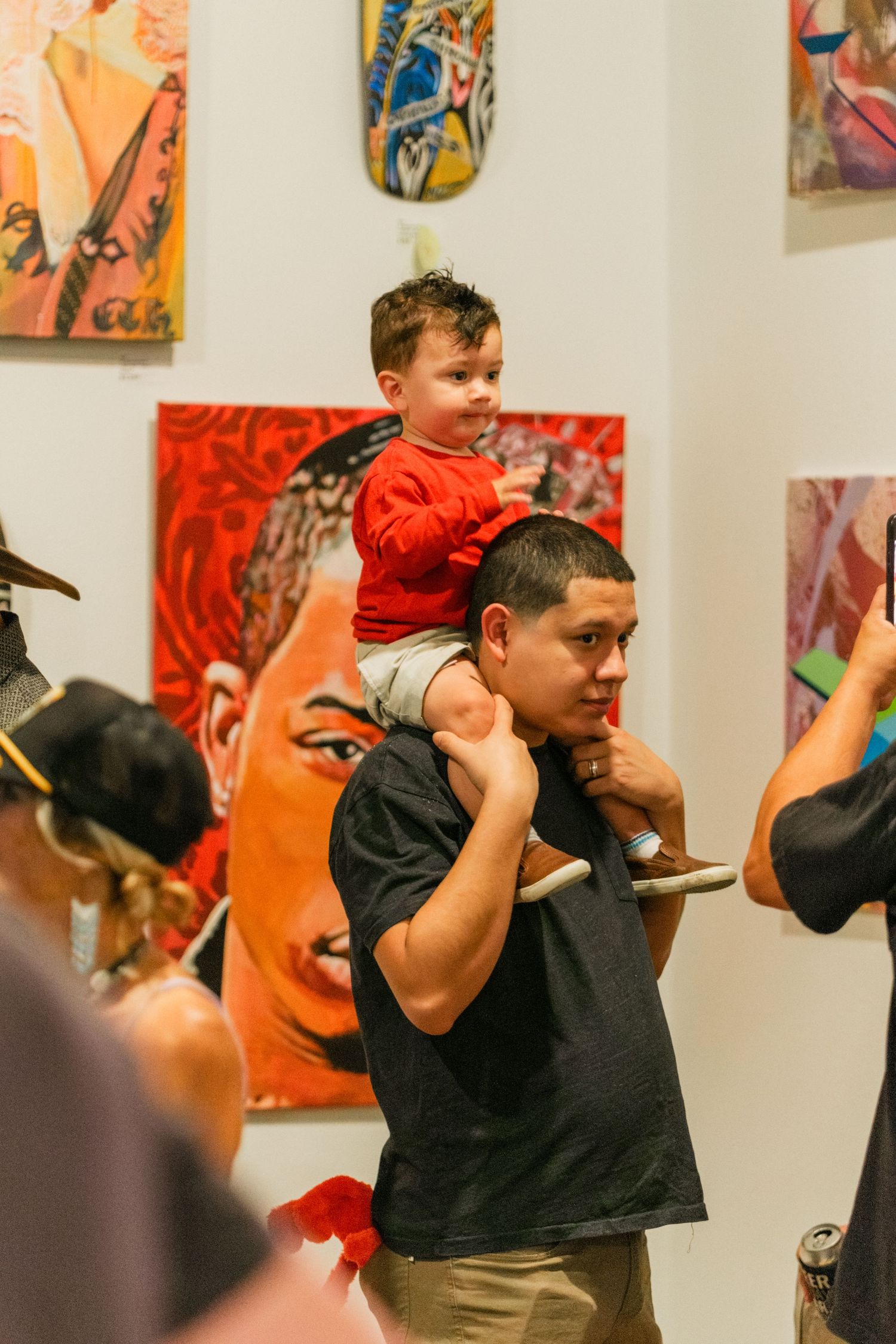 Father carrying young child on shoulders while viewing vibrant paintings at a crowded art gallery opening.
