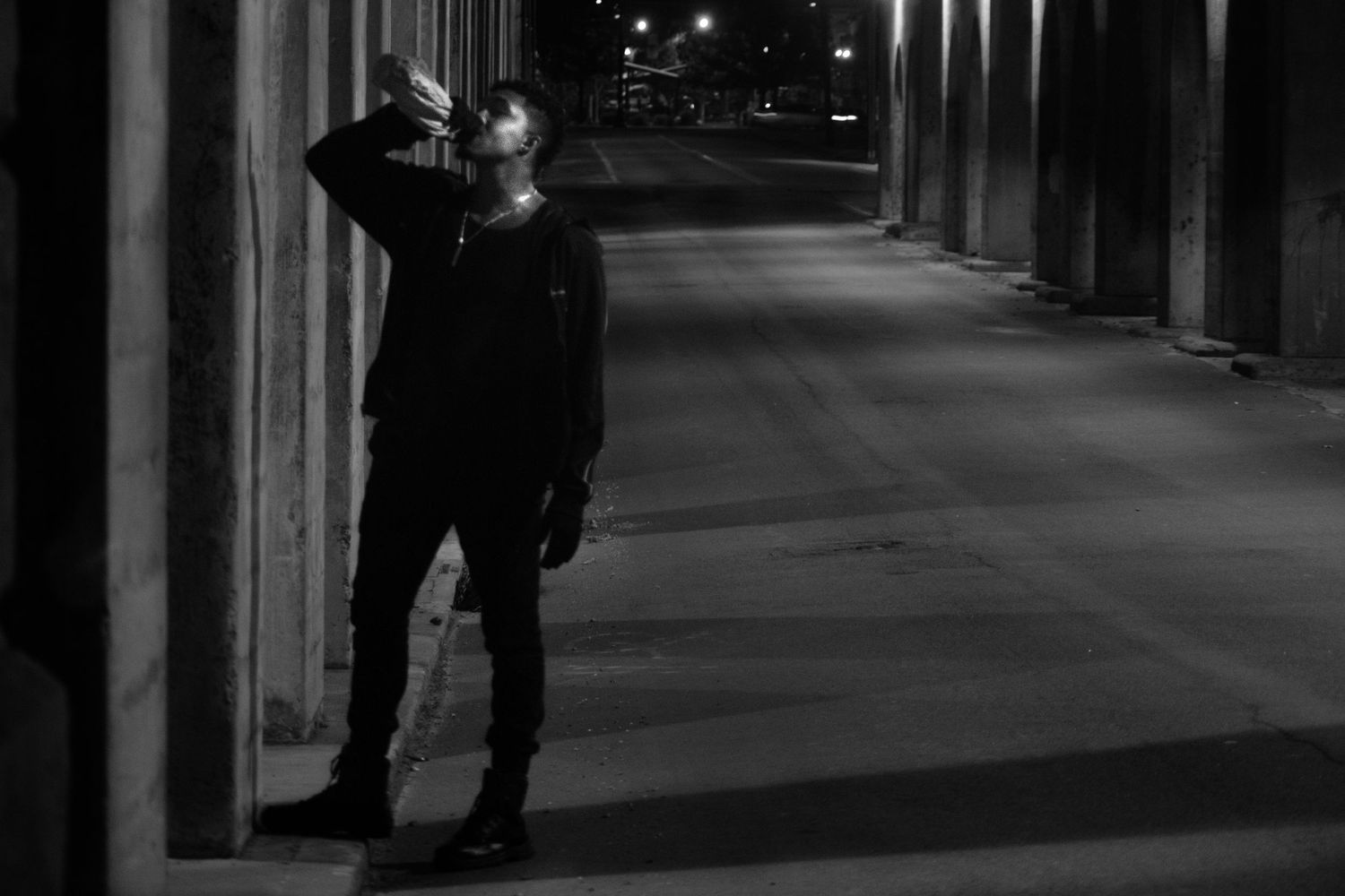 Black and white night photo of a person leaning against a column on a dark urban street.