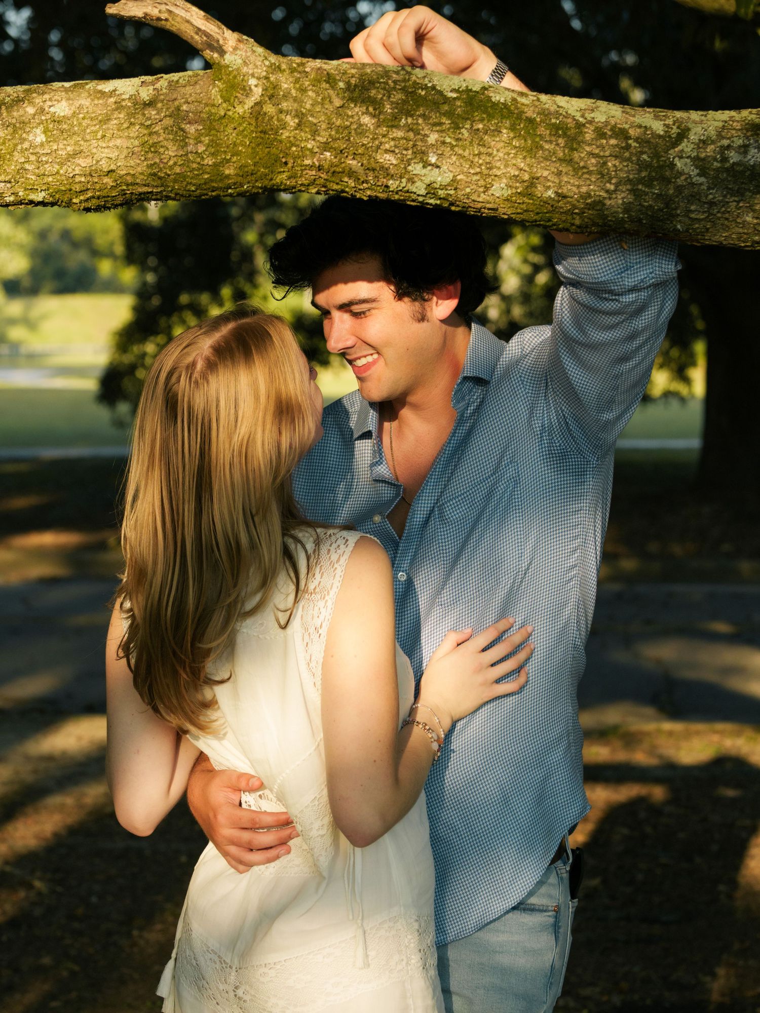 Romantic couple embracing under a tree branch in a sun-drenched park setting.