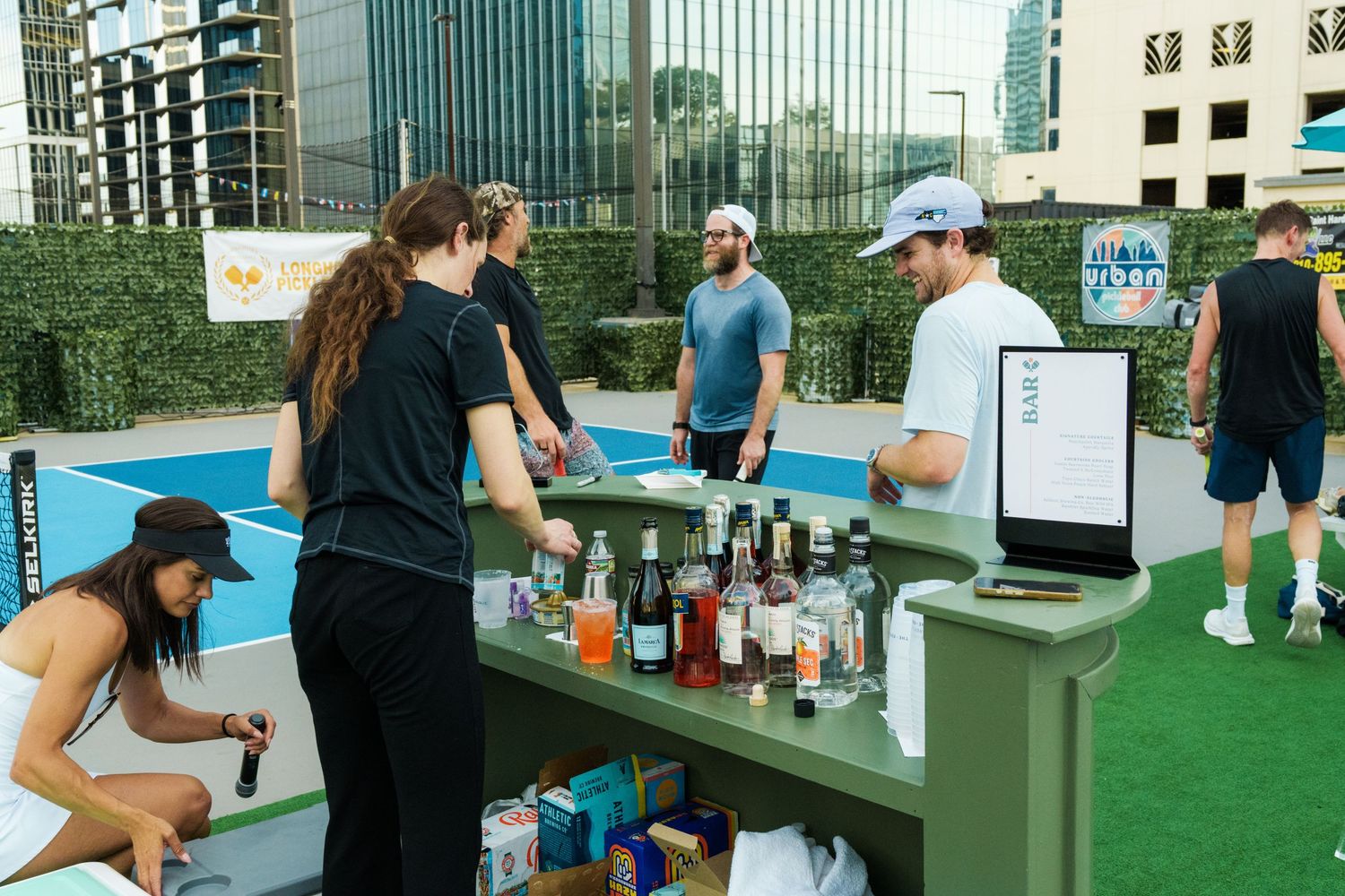 Rooftop bar setup near a pickleball court in an urban setting with guests browsing drinks and bottles.