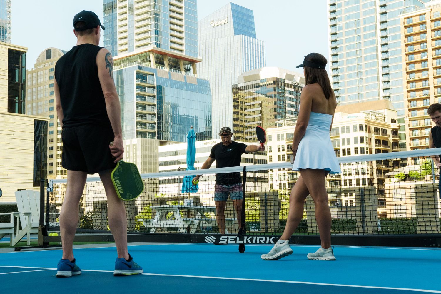 Players on a rooftop pickleball court with Selkirk paddles, city skyline backdrop on a sunny day.