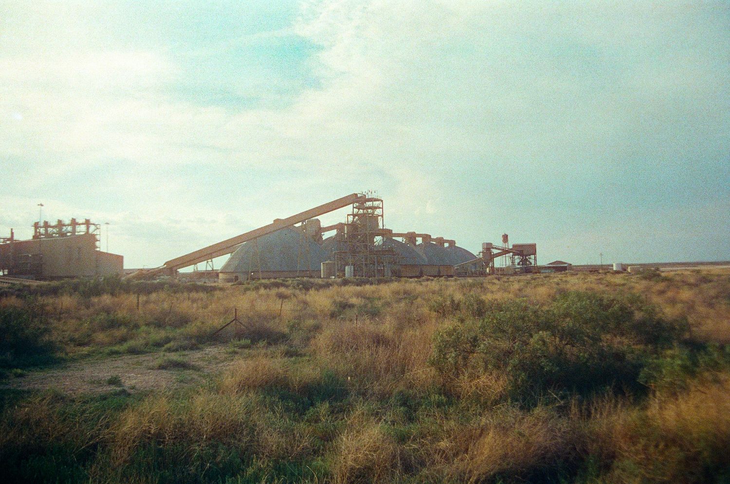 Abandoned industrial mining facility with conveyor belts and machinery surrounded by dry grassland under a cloudy sky.