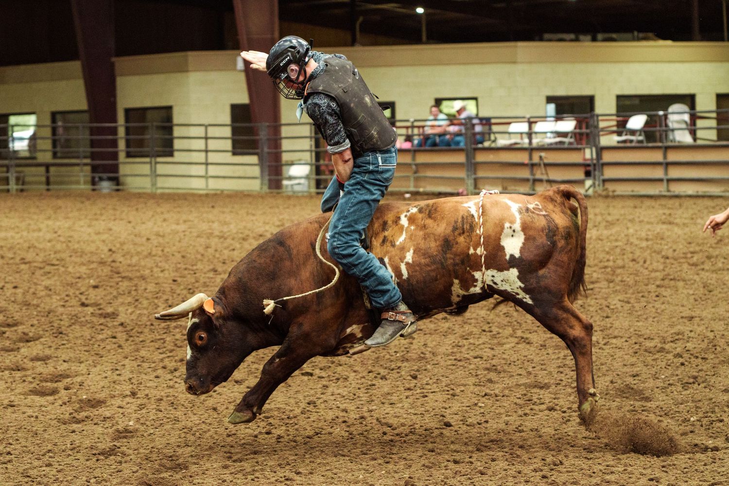 A bull rider in blue jeans grips a bucking brown bull during an indoor rodeo competition on a dirt arena floor.