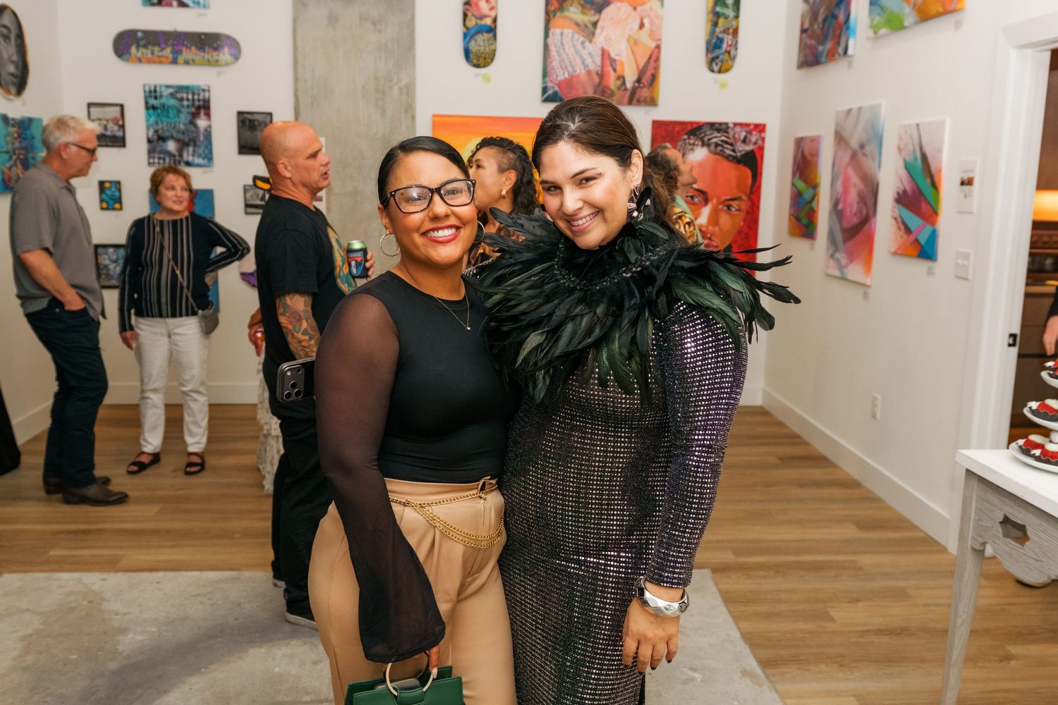Two women smiling together at an art gallery event, one wearing a feathered outfit.