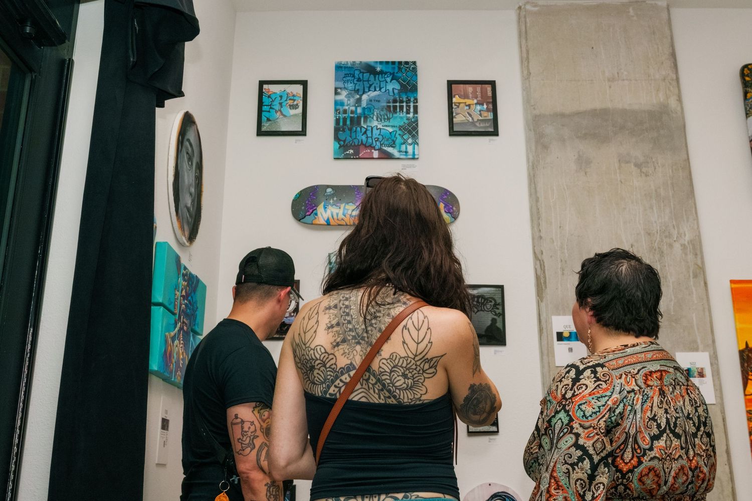 Three gallery visitors with tattoos view street art and graffiti paintings displayed on white gallery walls.