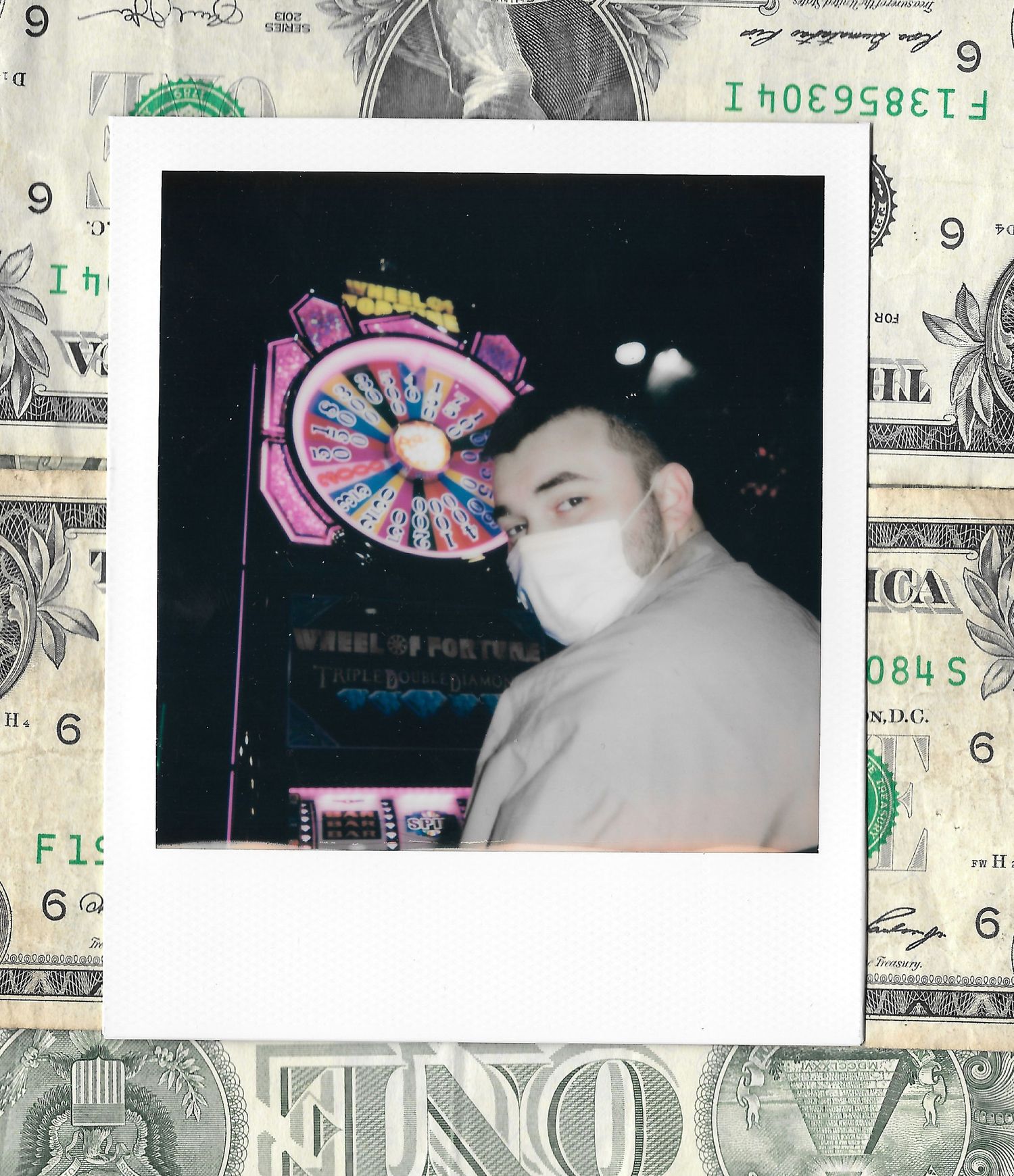 A person wearing a face mask poses in front of a neon Wheel of Fortune sign, framed as a Polaroid on dollar bills.