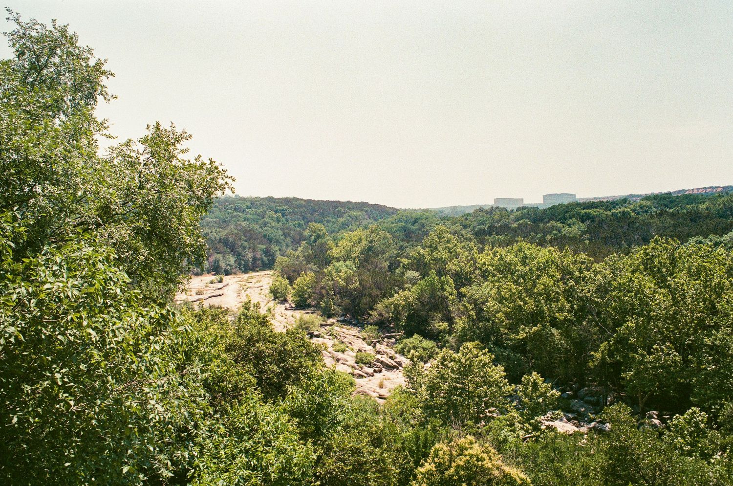 Scenic rocky canyon surrounded by lush green trees and forest with distant buildings visible on the horizon.