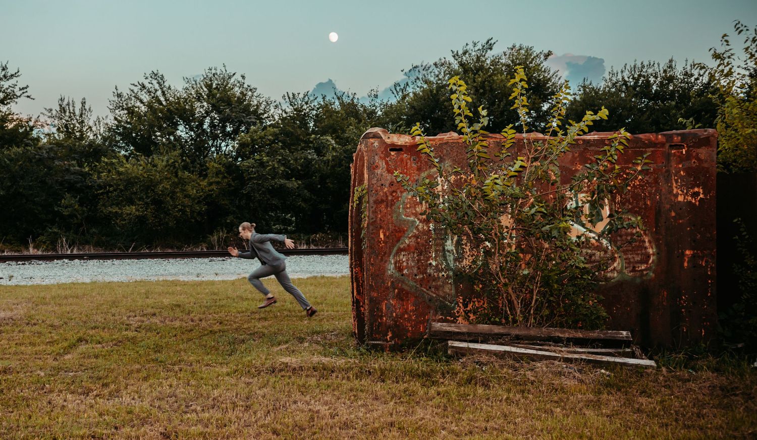 Person pushing a large rusty red container in an overgrown outdoor field at dusk.