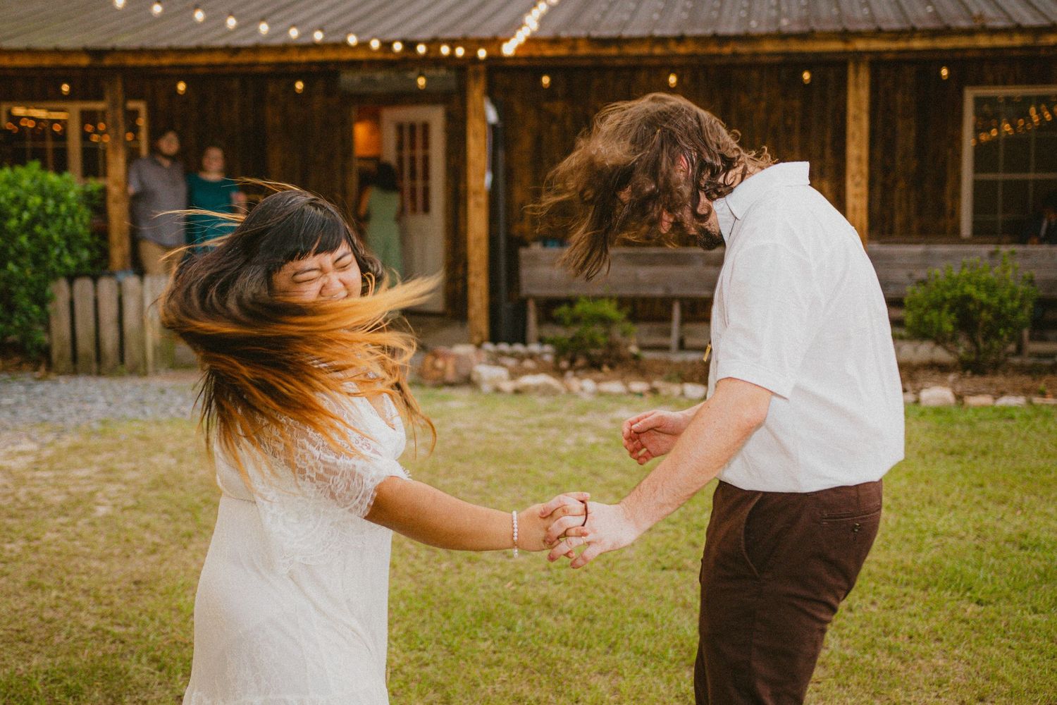 Joyful couple spinning and dancing outdoors at rustic barn wedding with string lights.