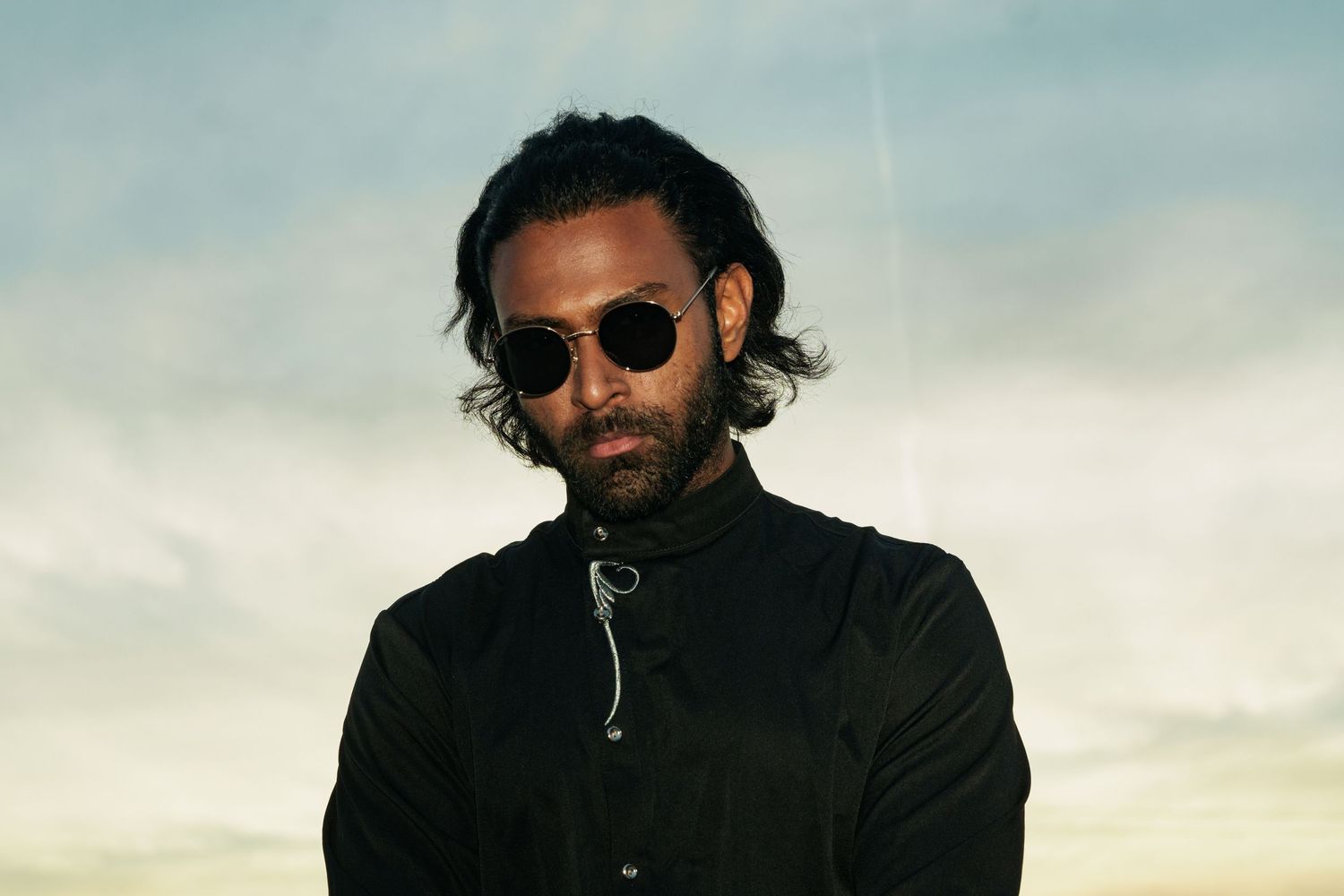 Man with long hair wearing black outfit and round sunglasses poses against dramatic cloudy sky backdrop.