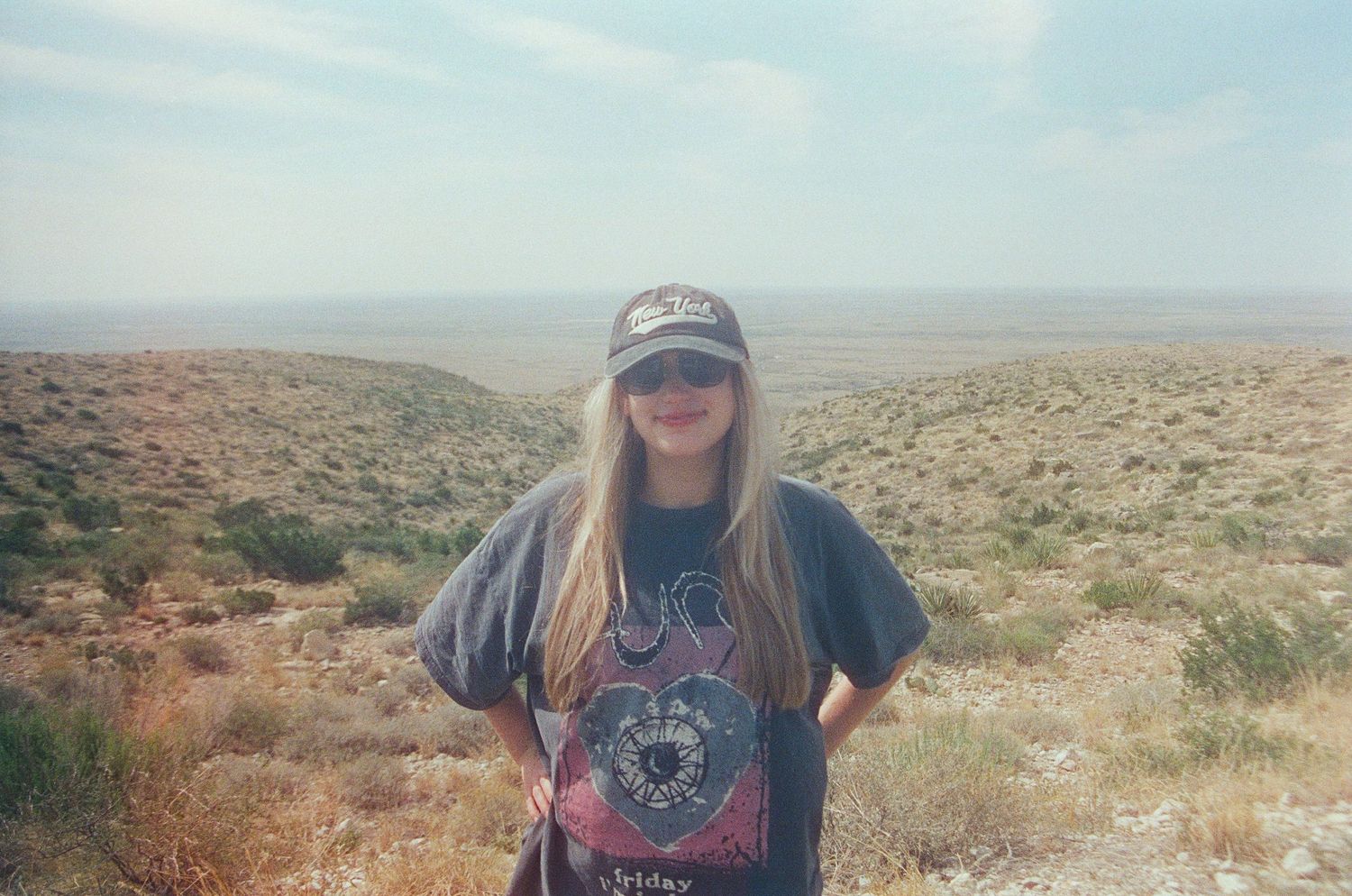 Young woman in graphic tee and cap stands before vast desert landscape under hazy sky.