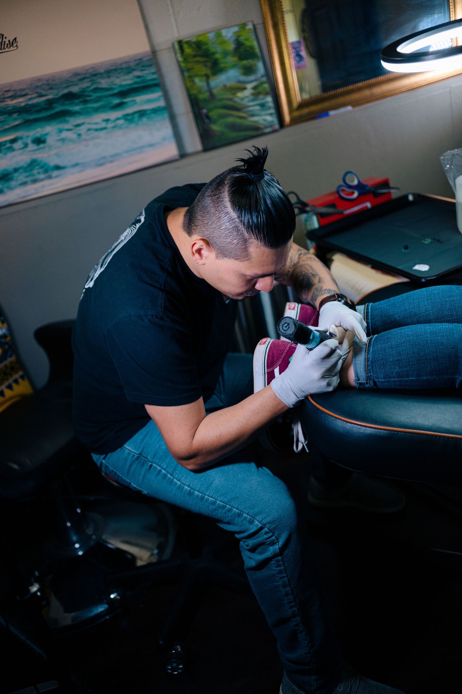 Tattoo artist in black shirt carefully tattooing a client's foot in a professional studio with artwork on walls.