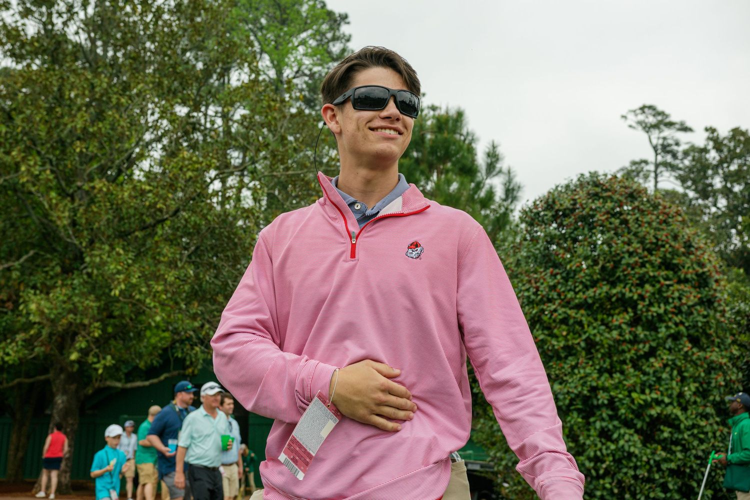 Young man in pink quarter-zip pullover and sunglasses smiling outdoors at a golf event.