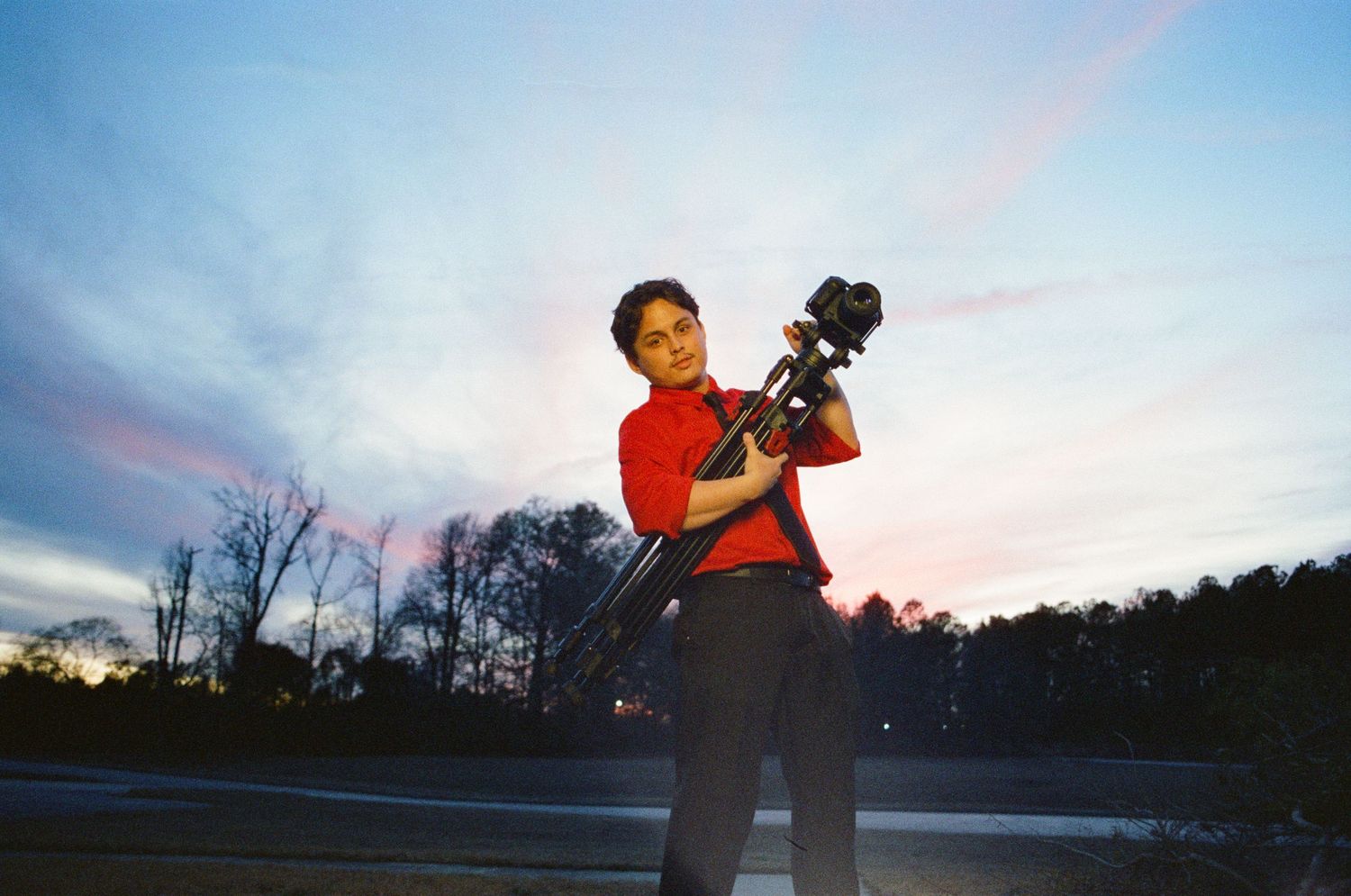 Person holding a camera on tripod outdoors at dusk with colorful sky behind.