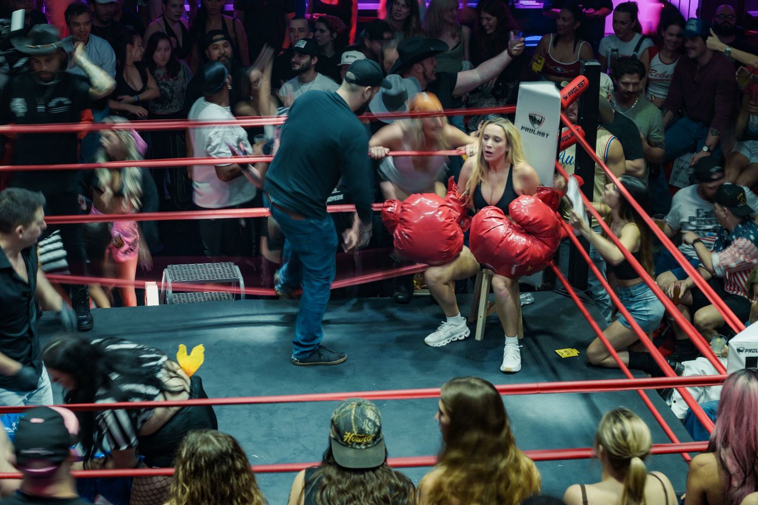A referee tends to a female boxer wearing red oversized gloves seated in the corner of a boxing ring.