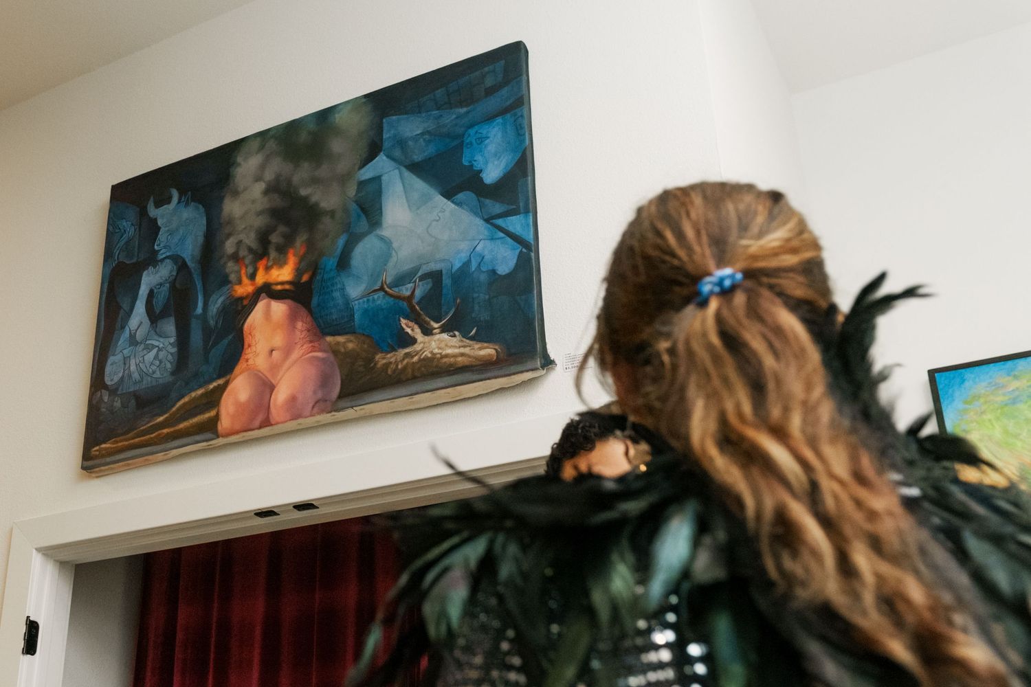 Gallery visitor with feathered accessories views a moody blue figurative painting on white wall.