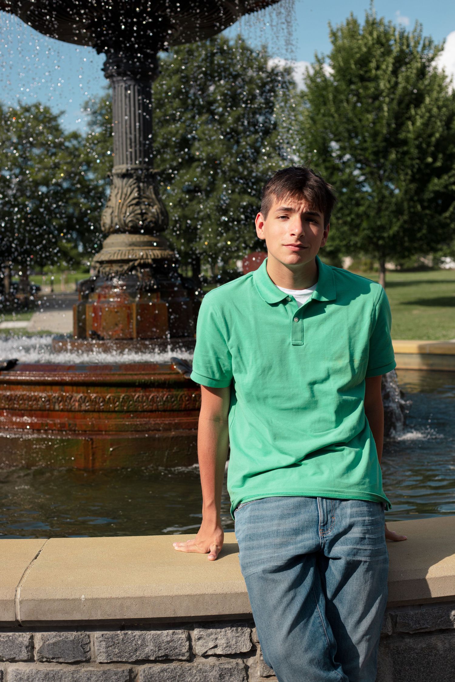 Teen boy in green polo shirt leaning against stone fountain in park.