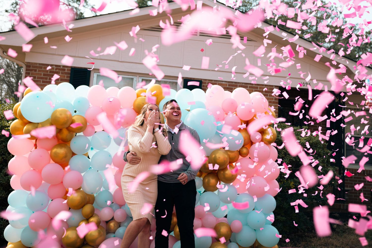 Couple celebrating gender reveal party with pink confetti and colorful balloon arch backdrop.
