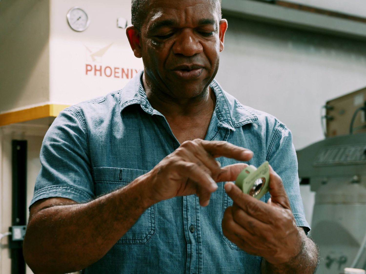 Factory worker carefully inspects a small green precision-machined component by hand.