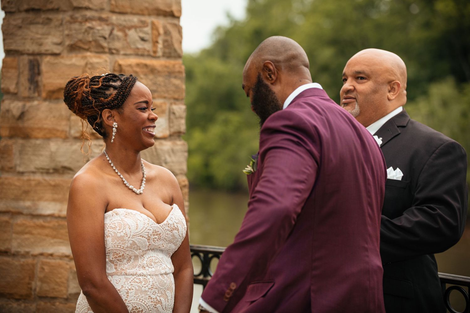 Bride in strapless lace gown smiling at groom in burgundy suit during outdoor wedding ceremony.