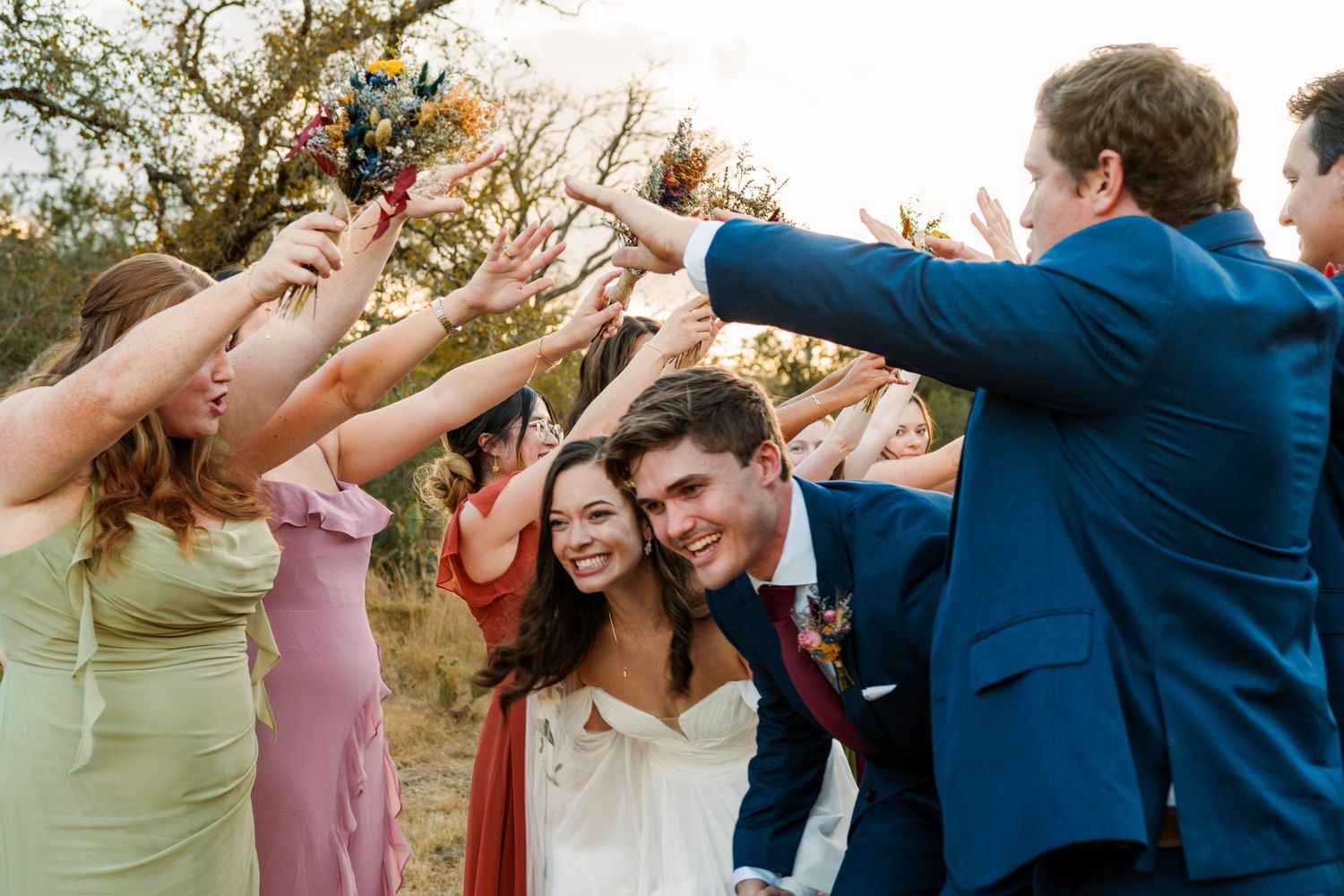 Bride and groom walk through a joyful tunnel of cheering wedding guests in colorful bridesmaid dresses outdoors.