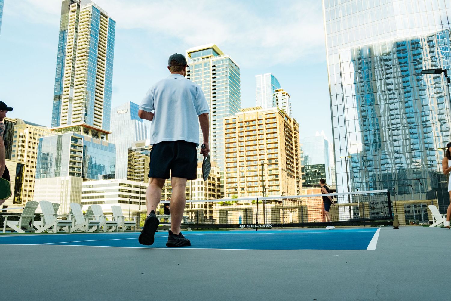 Man playing tennis on a rooftop court surrounded by modern city skyscrapers on a sunny day.