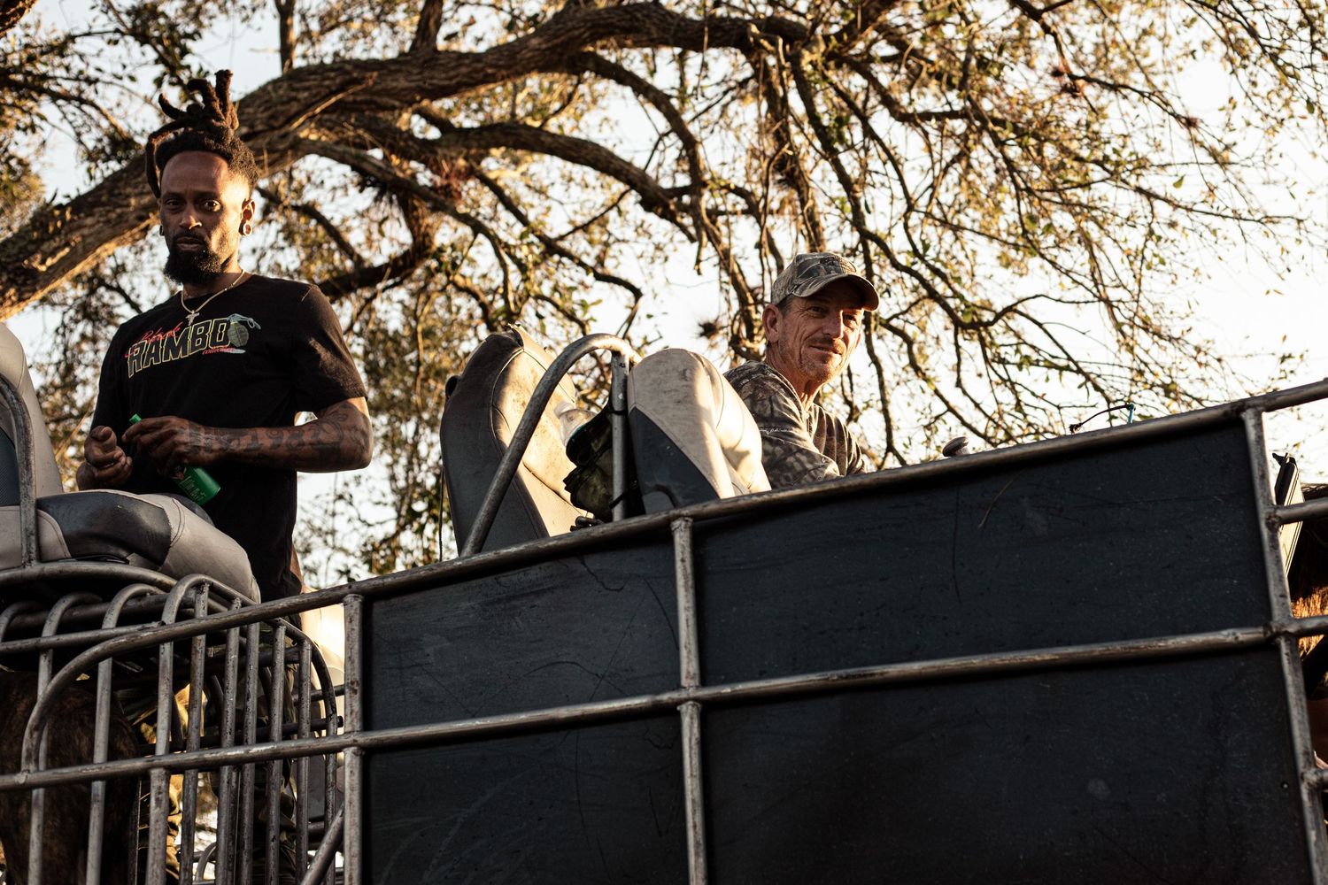 Two men standing on a flatbed trailer outdoors under trees at a daytime event.