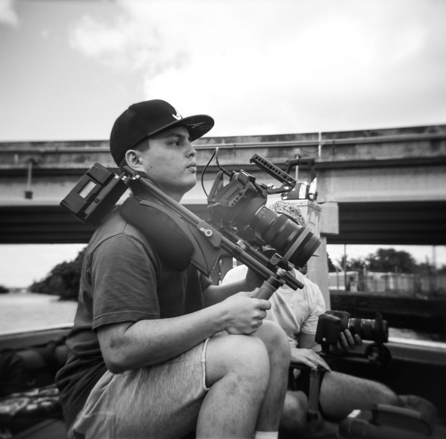 Black and white photo of a cameraman holding a professional film camera outdoors near a bridge.