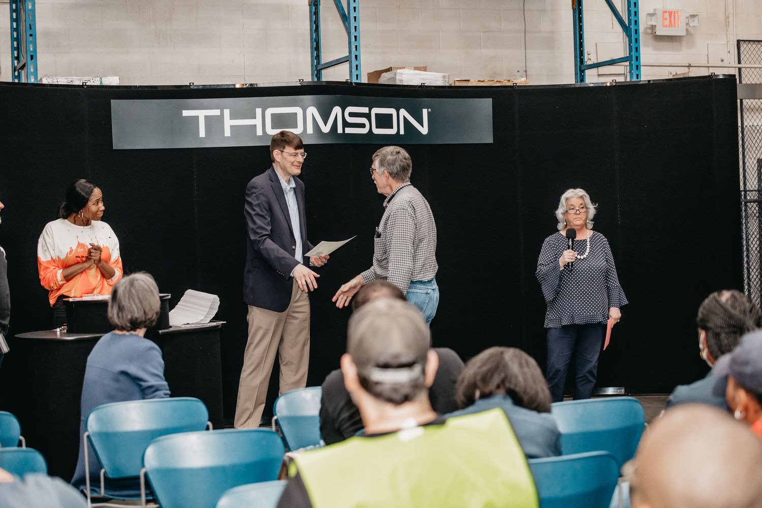 Professionals exchanging documents at a Thomson corporate event in a warehouse setting with seated audience.