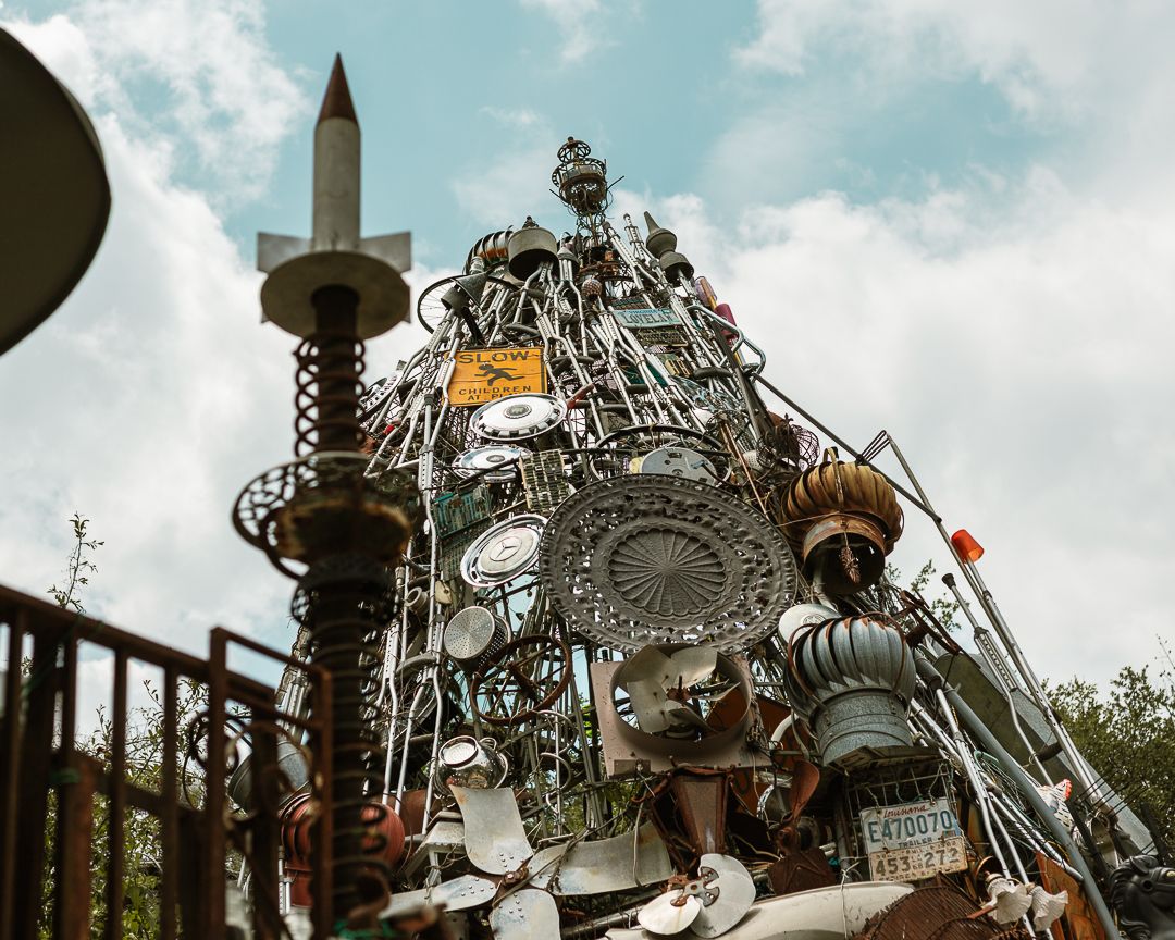 Eclectic outdoor sculpture made of salvaged metal parts, hubcaps, and a rocket, under a partly cloudy sky.