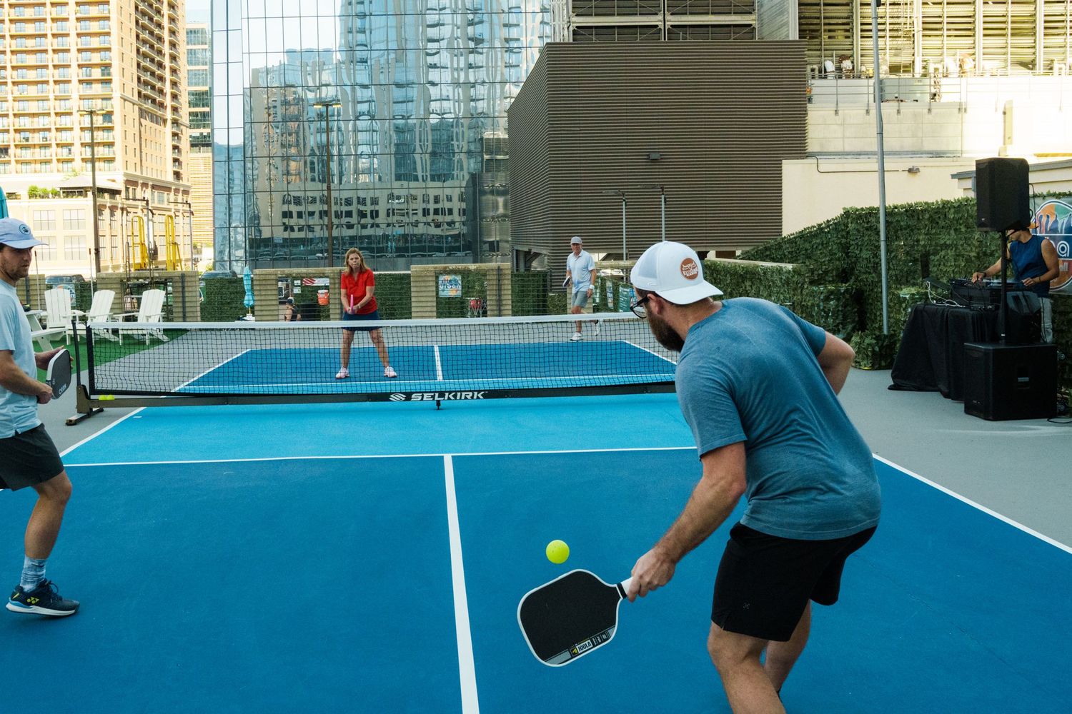 Players compete in a pickleball match on a rooftop court with city skyline views on a sunny day.