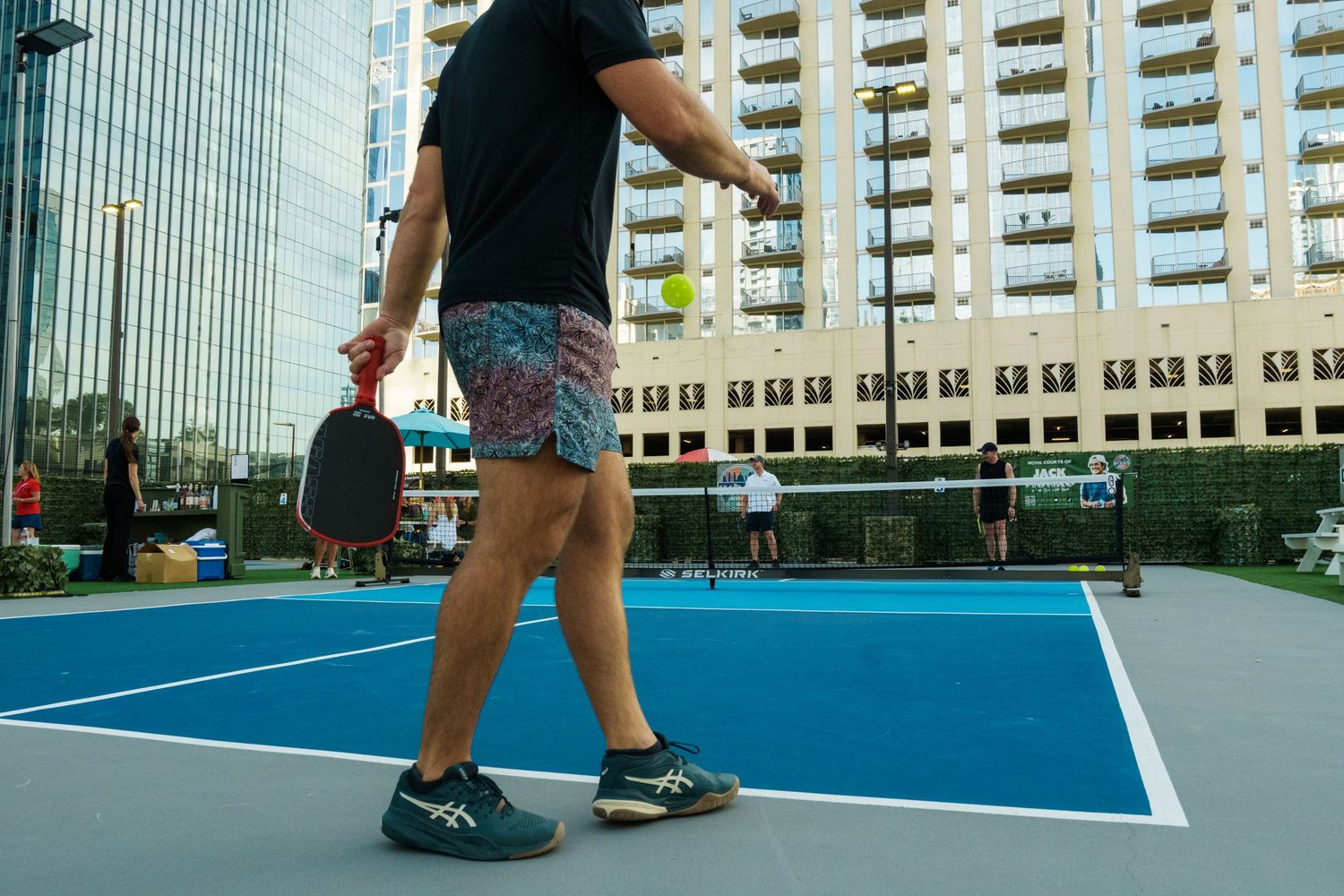Player holding a pickleball paddle on a blue outdoor court with urban buildings in the background.