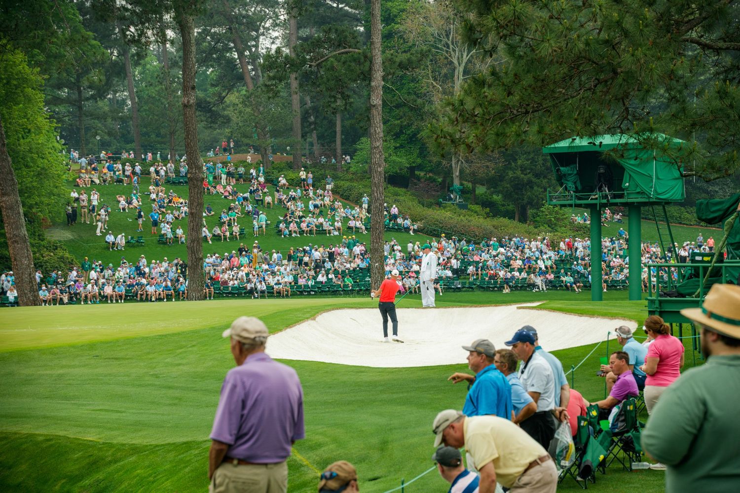 Golfers and spectators gather around a sand bunker on a lush green golf course during a major tournament.