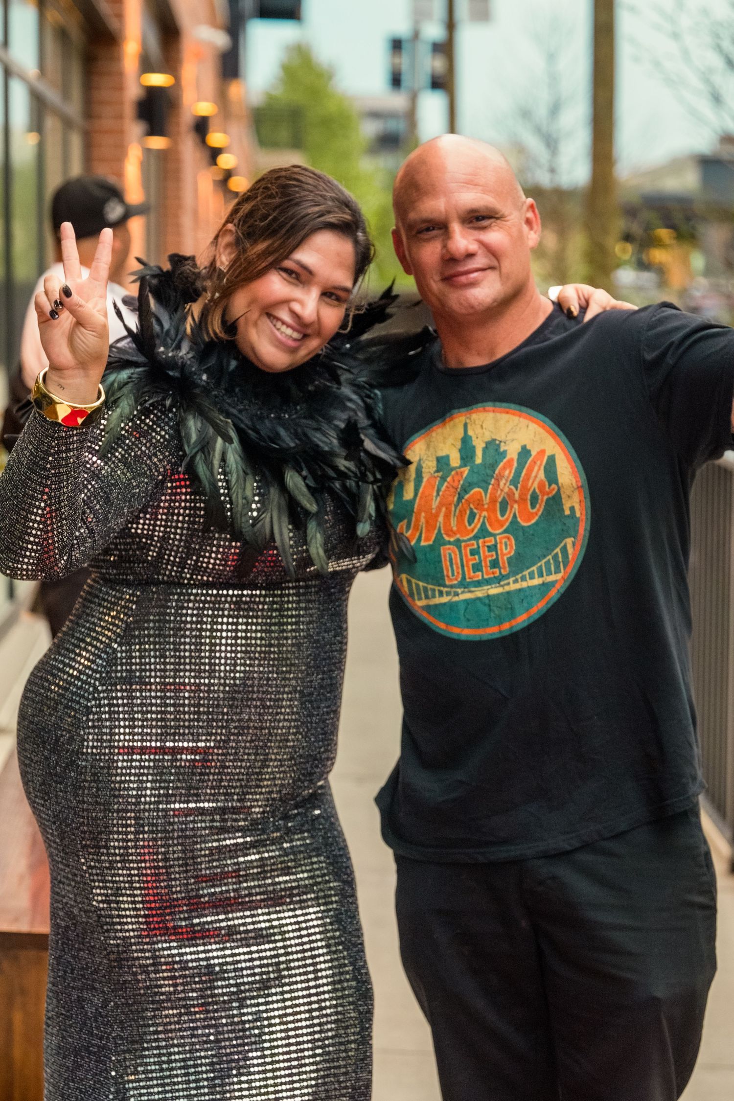 Woman in sequin dress and man in Mobb Deep t-shirt posing together outdoors at a street event.