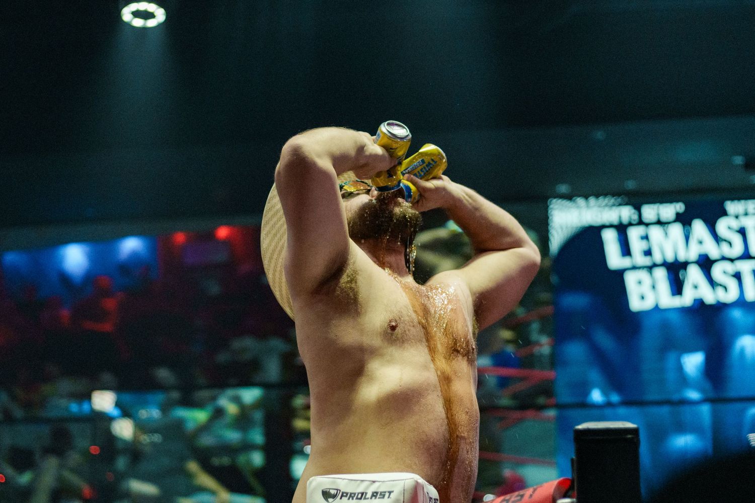 Shirtless boxer holds championship belts above his head in a boxing ring under blue arena lights.