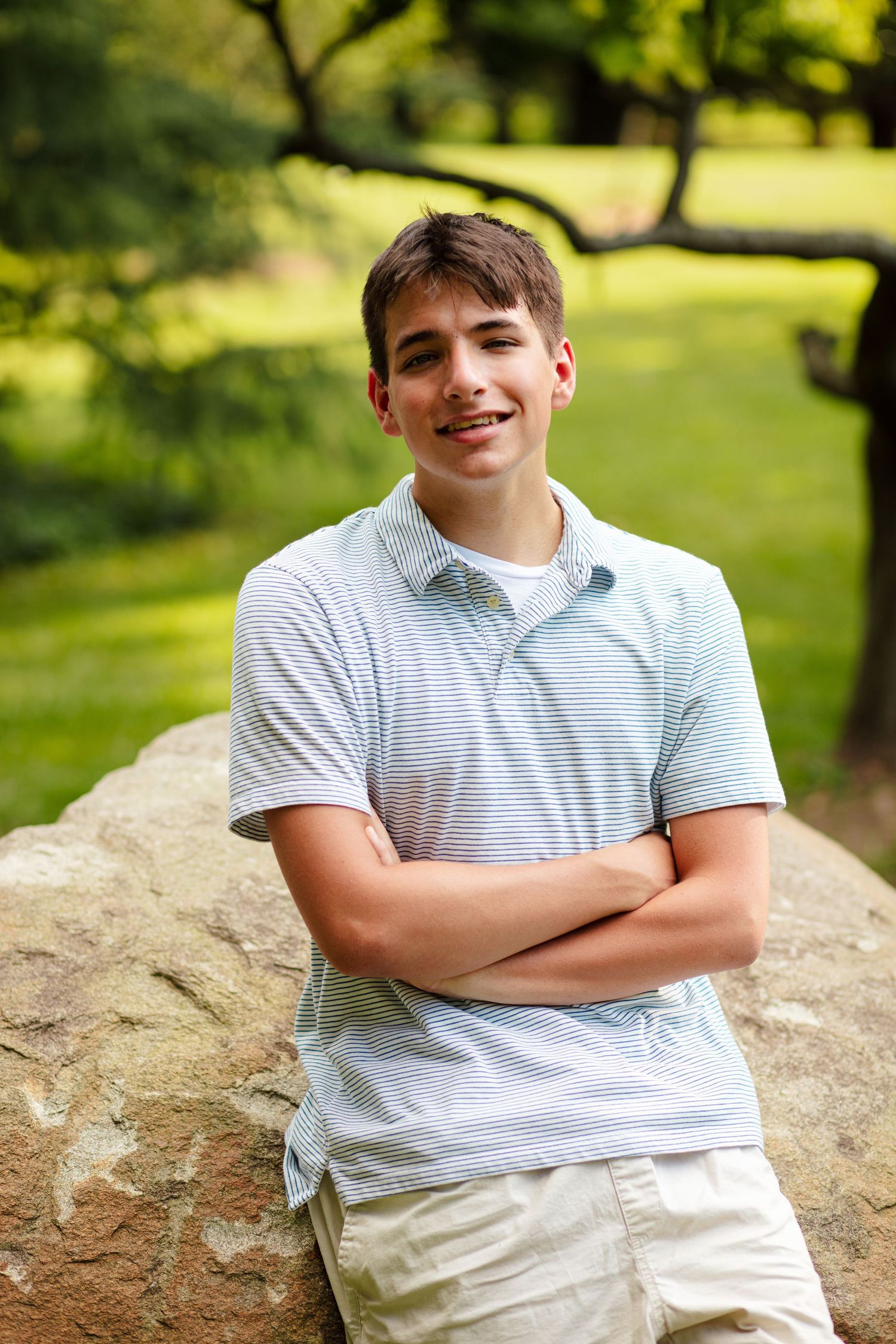 Teen boy in striped polo shirt leaning against boulder with arms crossed outdoors.
