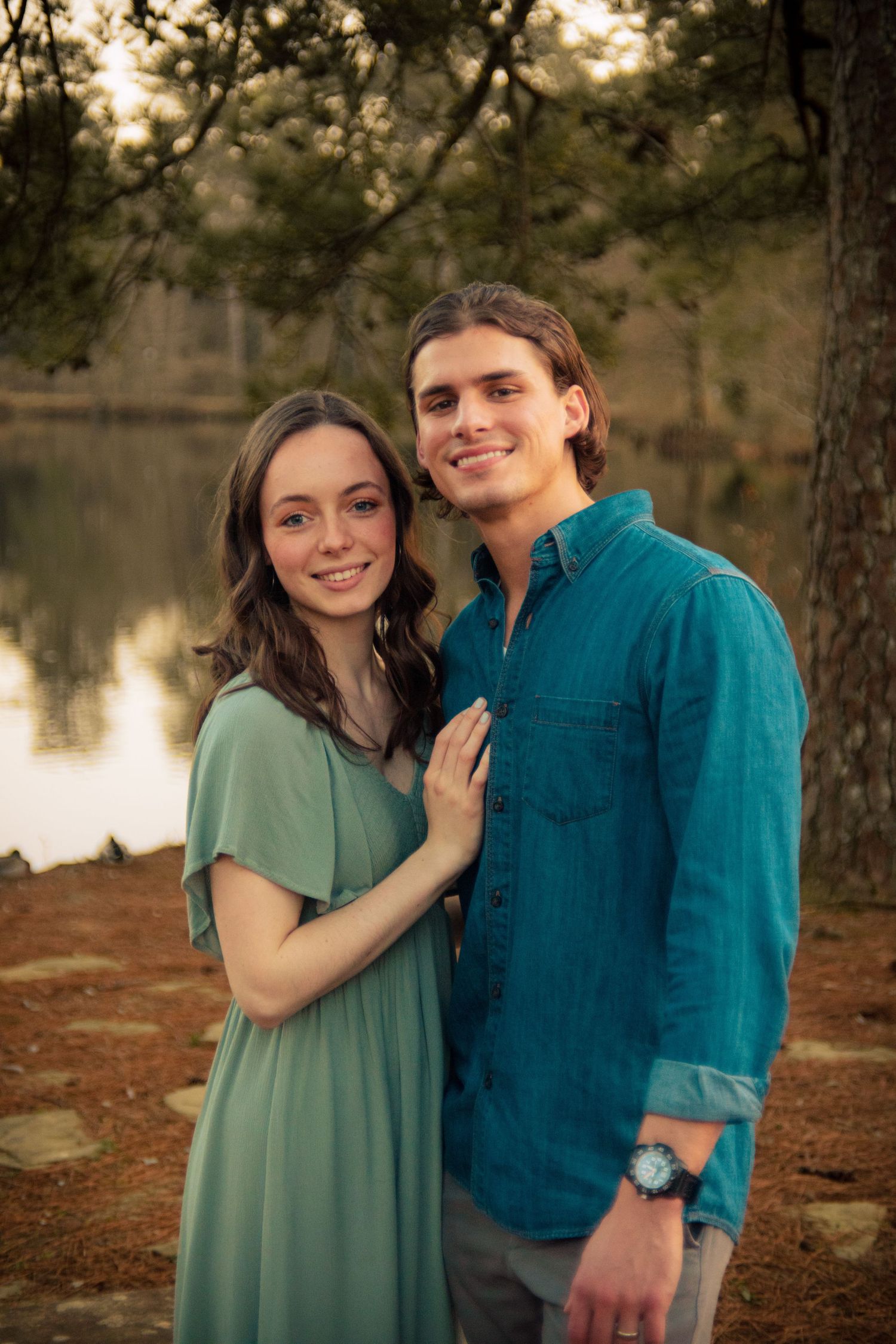Smiling young couple poses together near a serene lake surrounded by pine trees at dusk.