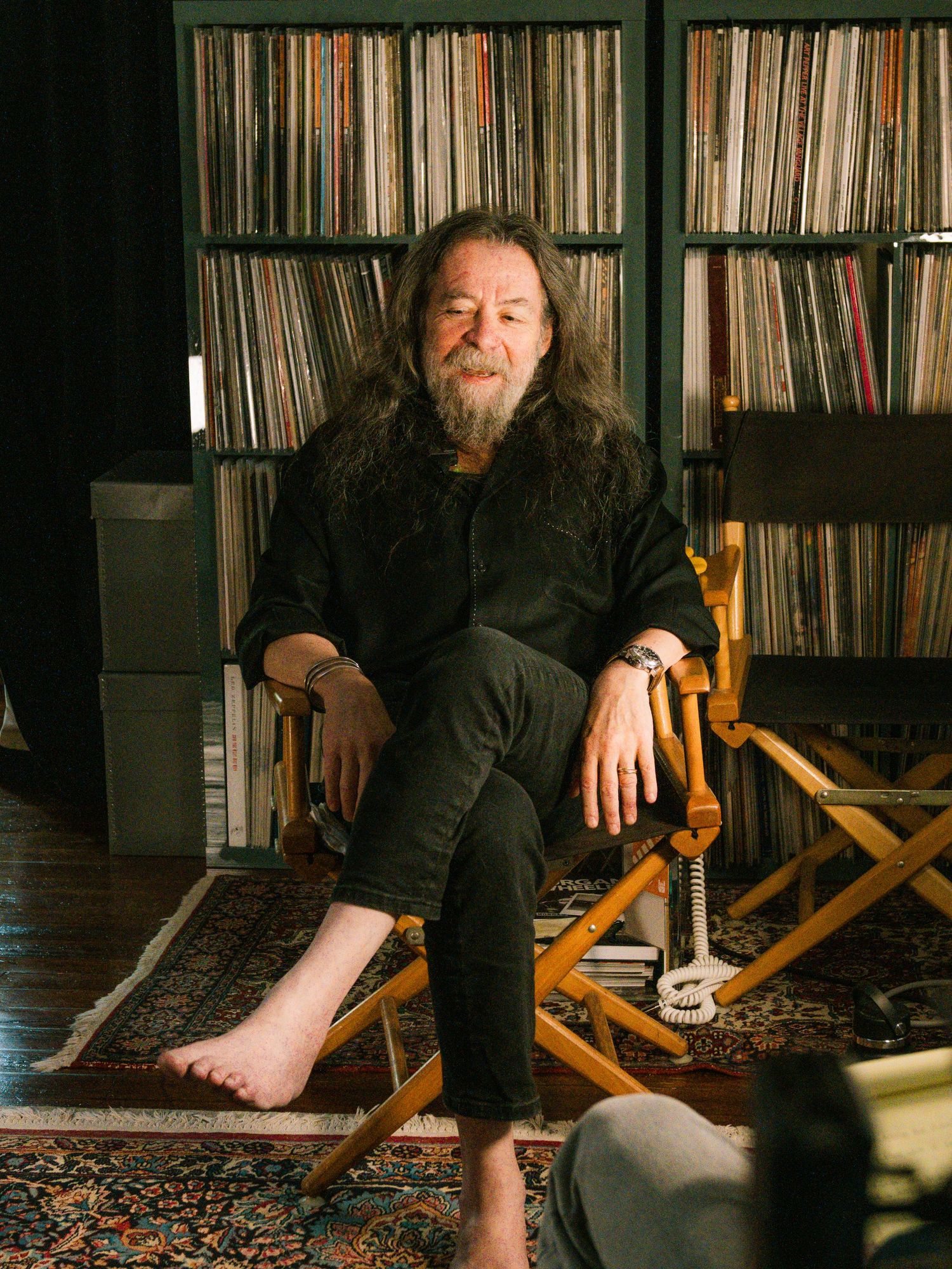 Long-haired bearded man sitting barefoot in director chair surrounded by vinyl record collection.