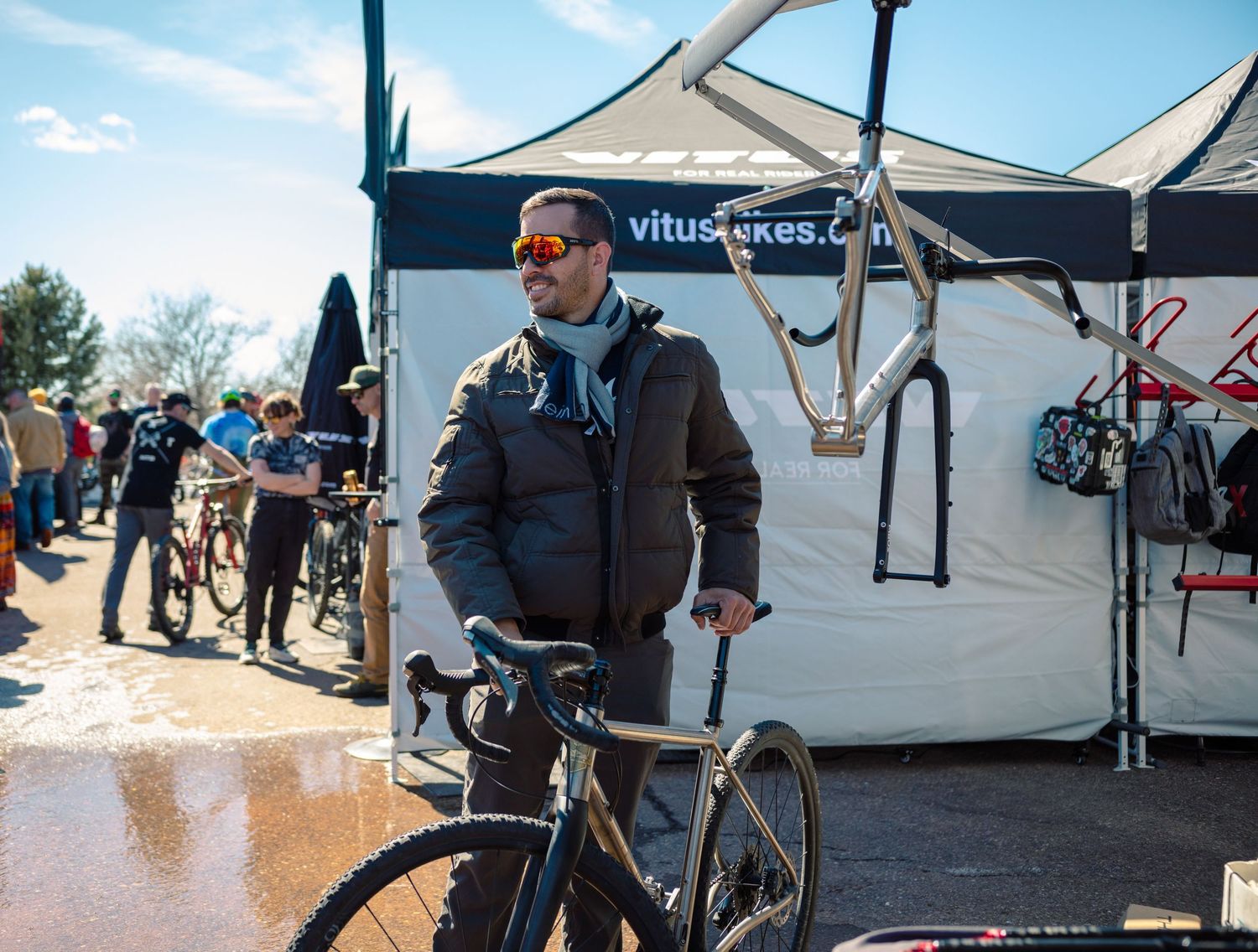 Man in sunglasses and brown jacket holds a black road bike at a Vitus Bikes outdoor cycling expo.