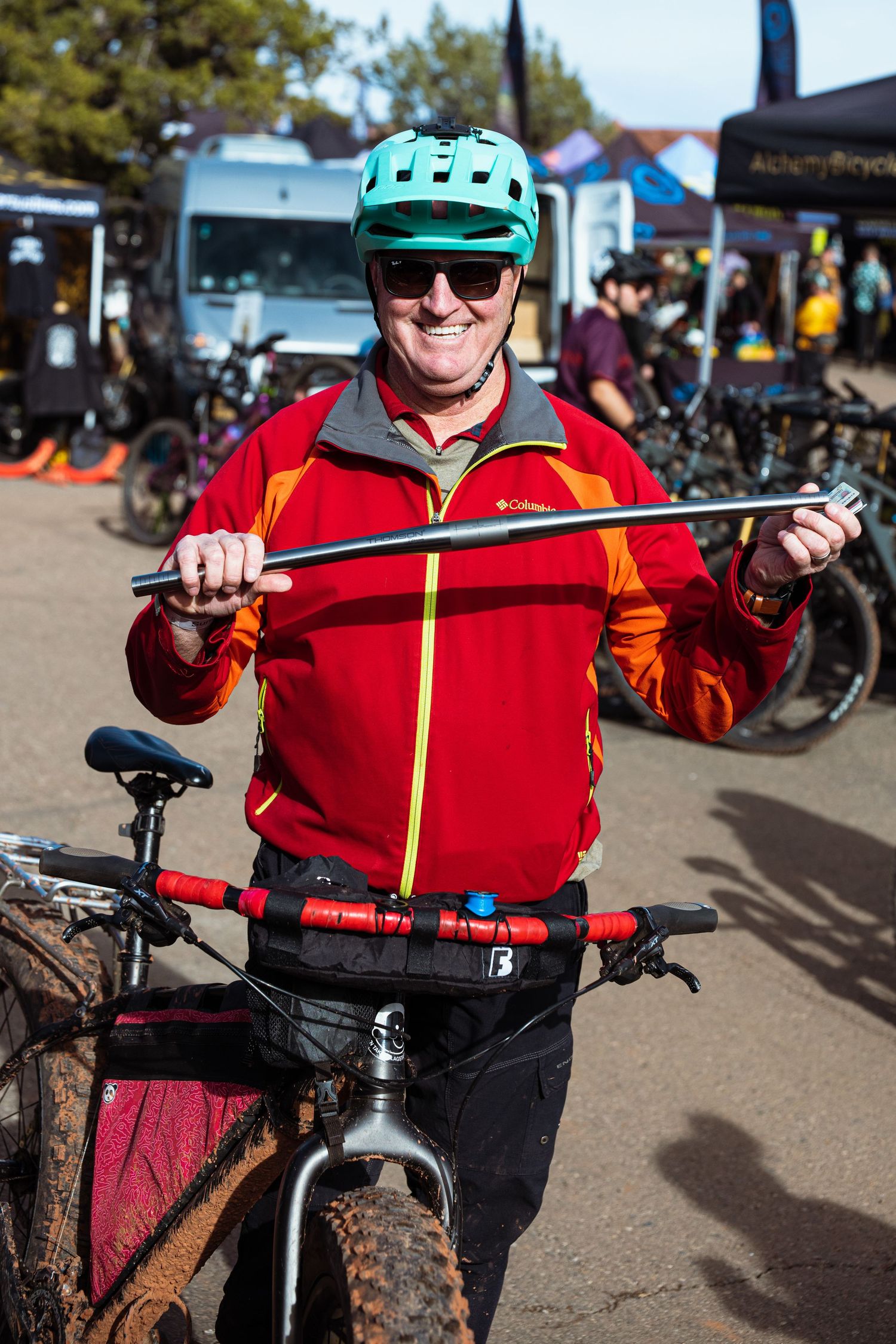 Smiling man in red Columbia jacket and teal helmet holding a metal bar next to a mountain bike.