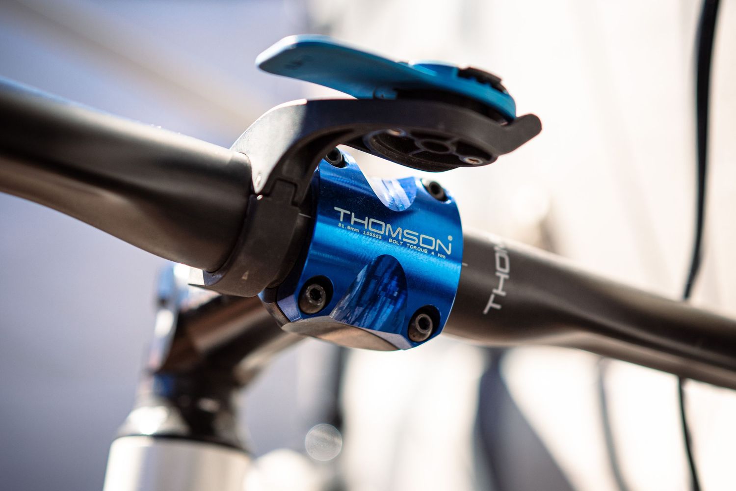 Blue Thomson stem with phone mount attached to a carbon fiber bicycle handlebar close-up detail shot.