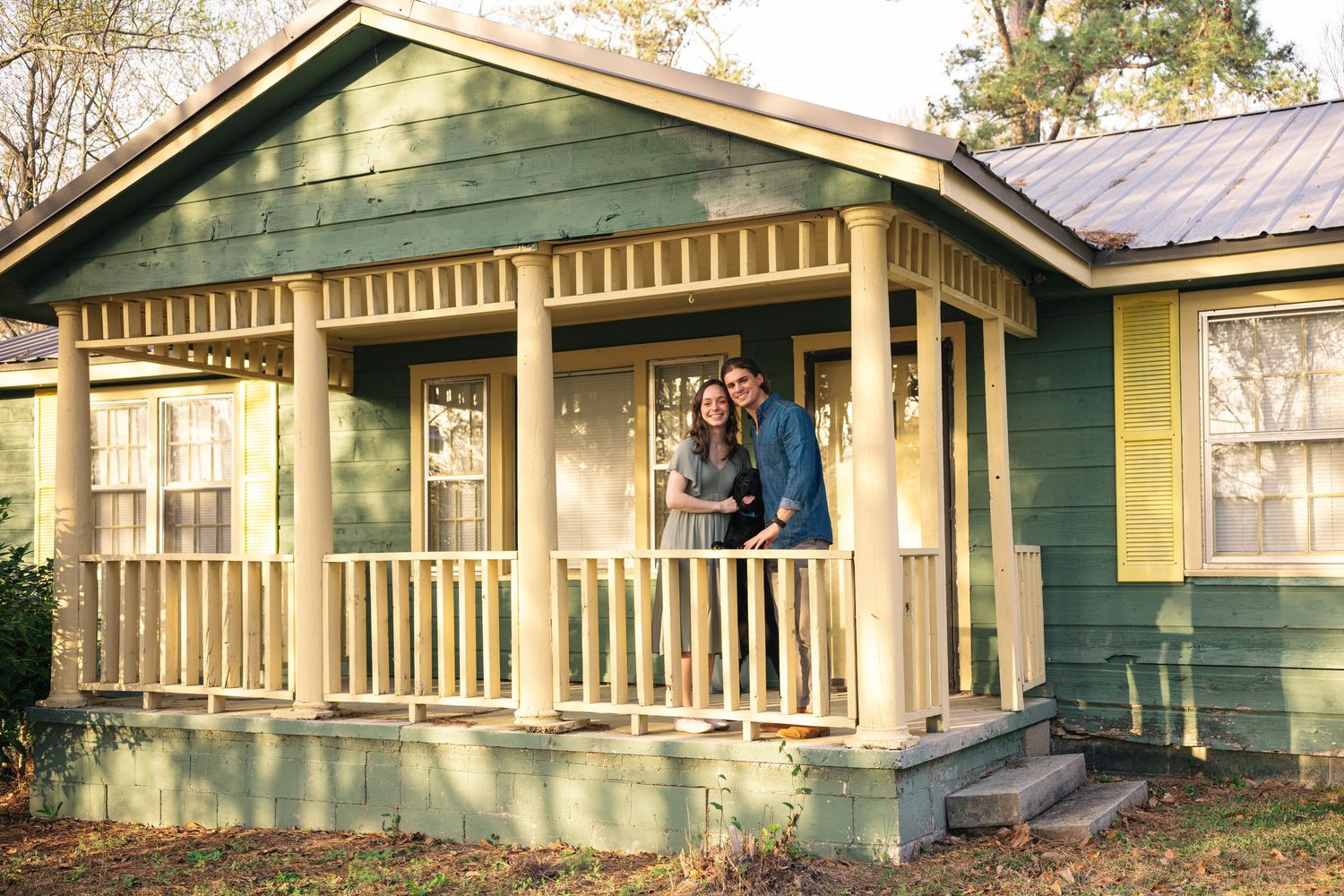 Smiling couple posing together on the porch of a charming green cottage home.